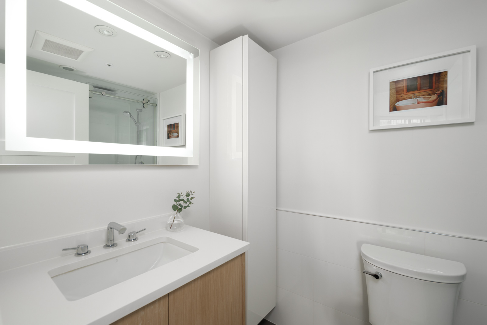 Modern bathroom with a white countertop sink, faucet, toilet, wall cabinet, illuminated mirror, and framed artwork on a white wall.
