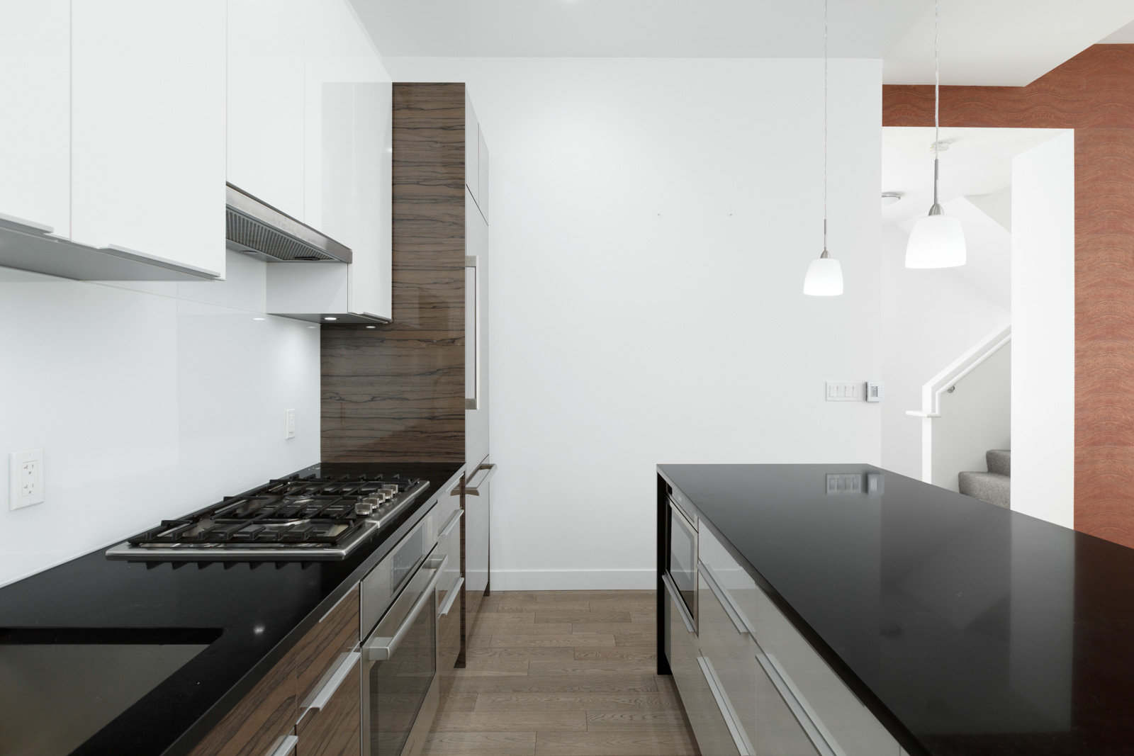 Modern kitchen with black countertops, gas stovetop, white cabinets, wooden accents, pendant lights, and a view of stairs in the background.