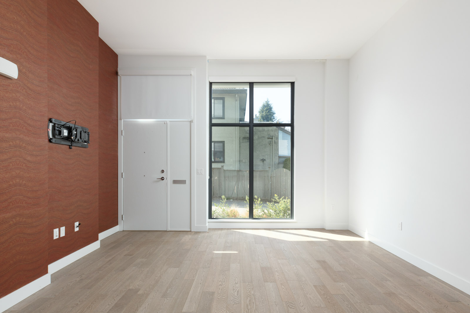 Empty modern room with light wood flooring, a large window, white walls, a red accent wall, and a door with transom window. TV mount attached to the red wall.
