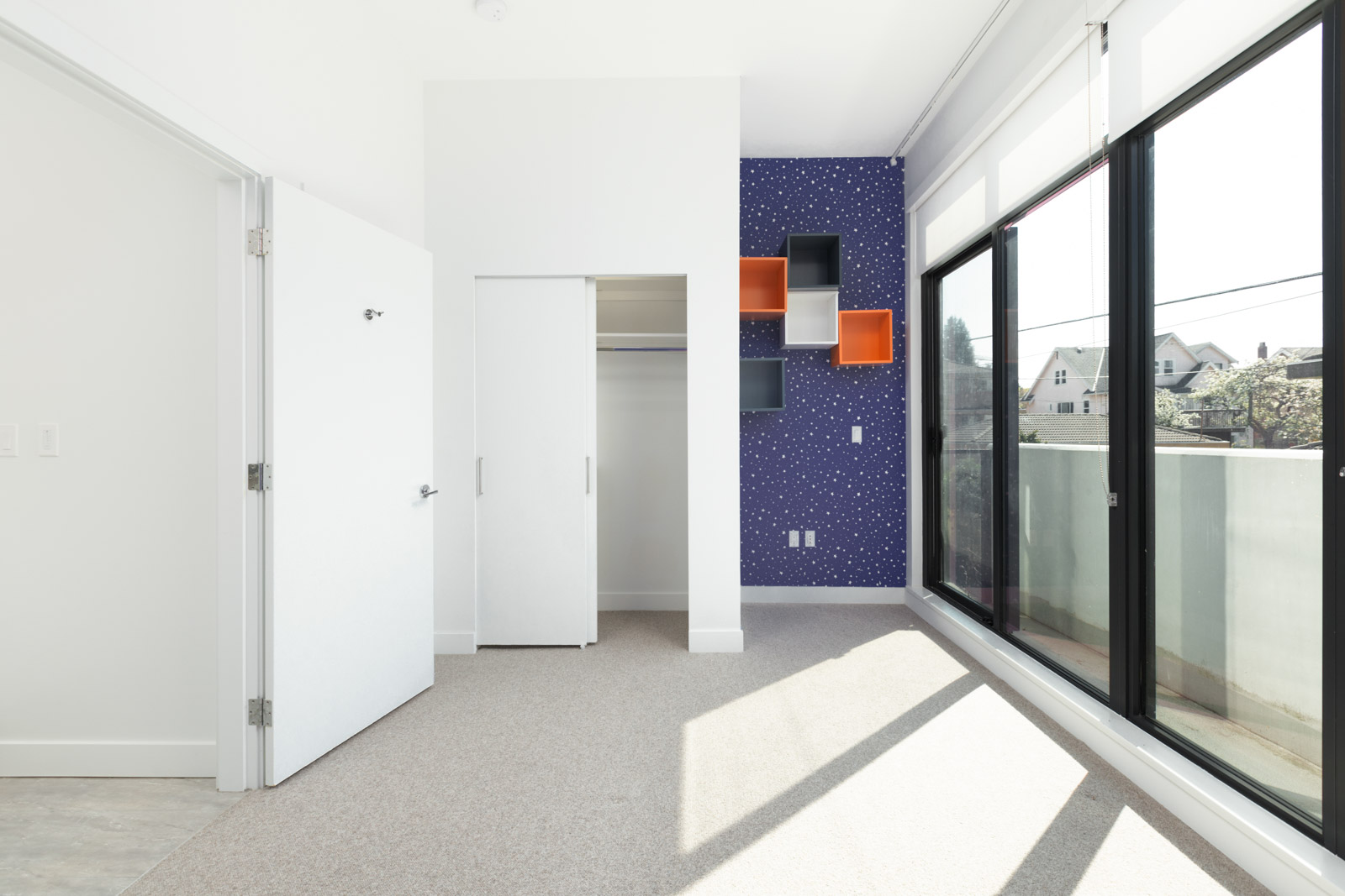 A bright, empty room with beige carpet, a closet, geometric wall shelves, large windows, and a blue accent wall with white dots.