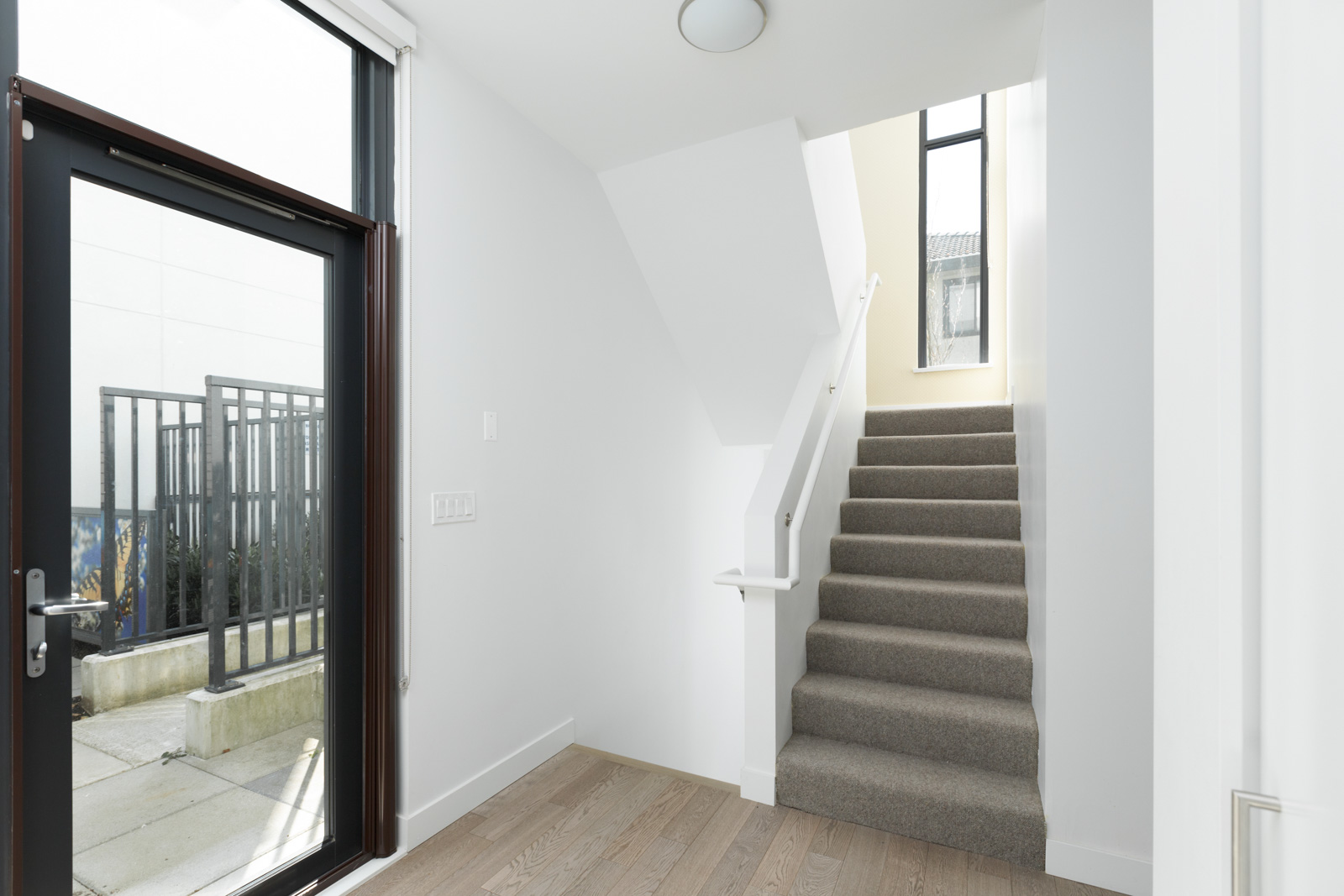 A modern interior entryway with a glass door, light wood floor, and a carpeted staircase leading up to a sunlit window.