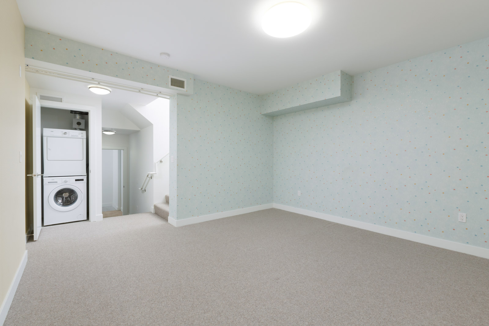 Empty carpeted room with light blue dotted wallpaper, a ceiling light, and a view of a hallway with a stacked washer and dryer near a staircase.