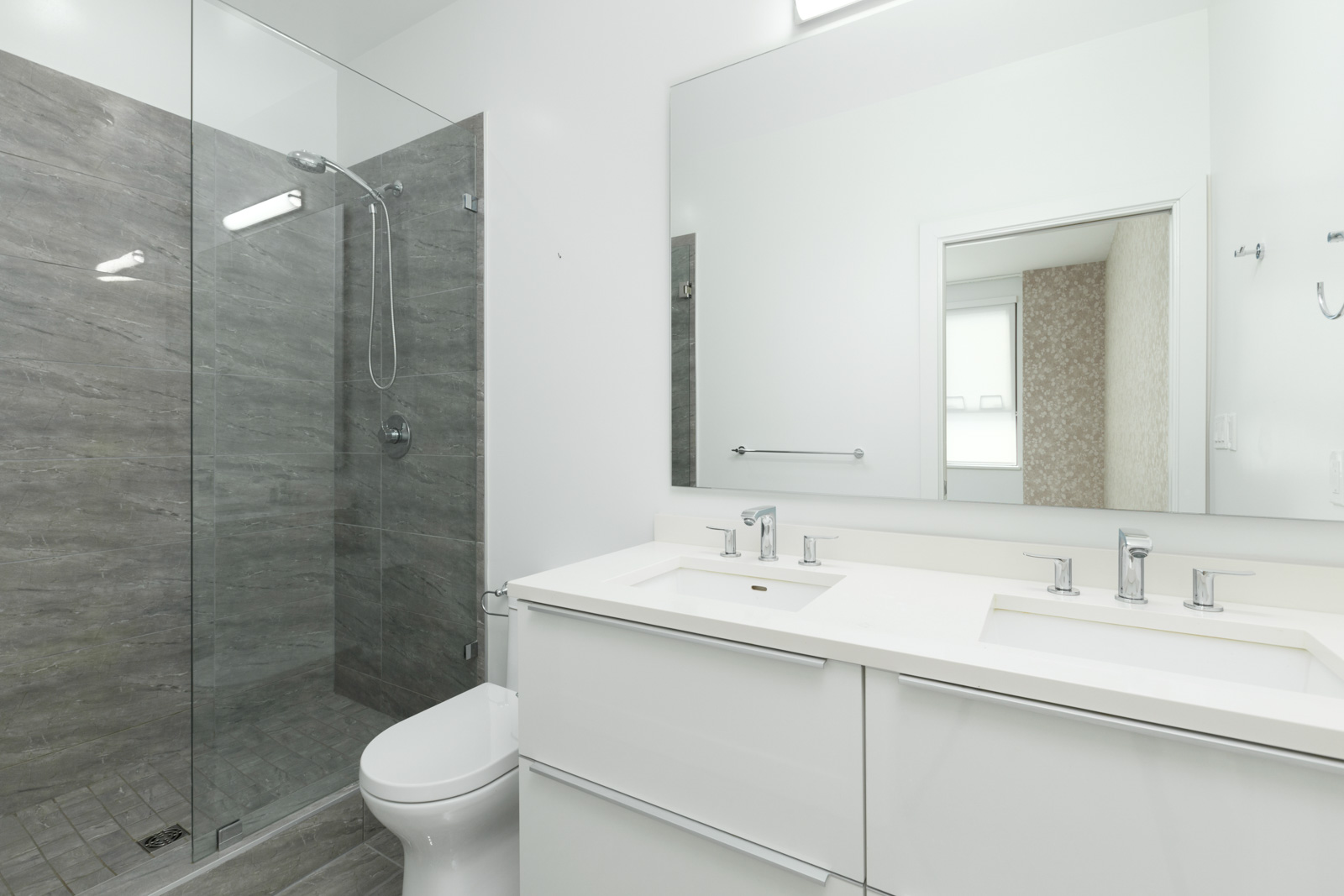 Modern bathroom with a glass-enclosed shower, gray tile walls, white double-sink vanity, large mirror, and a toilet.