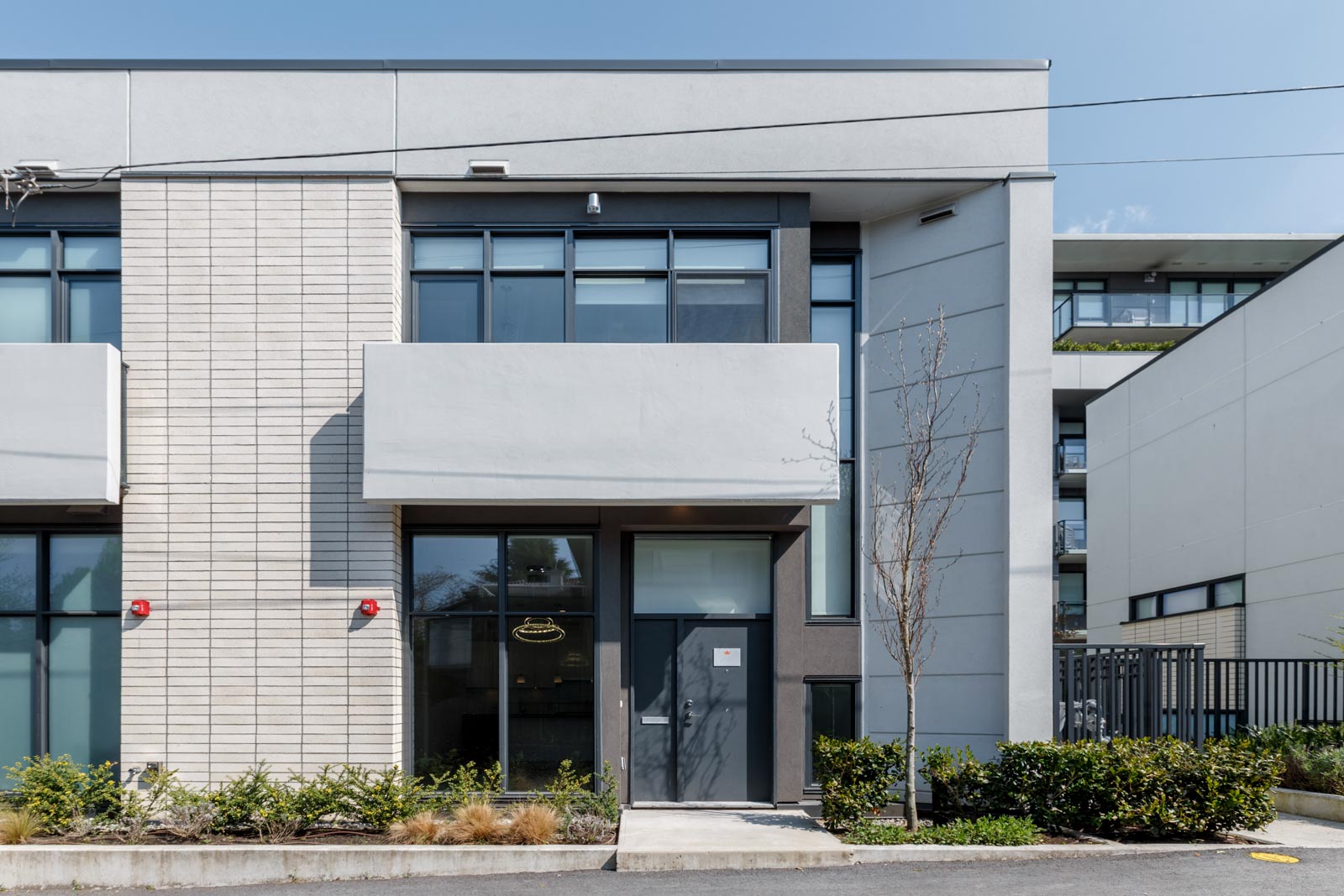Modern two-story building with large windows, a central entrance door, small balcony, and minimal landscaping in front.