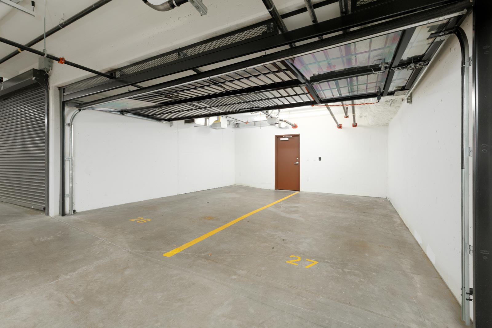Two empty parking spaces numbered 27 and 28 in a clean, well-lit, indoor parking garage with a brown door at the back.