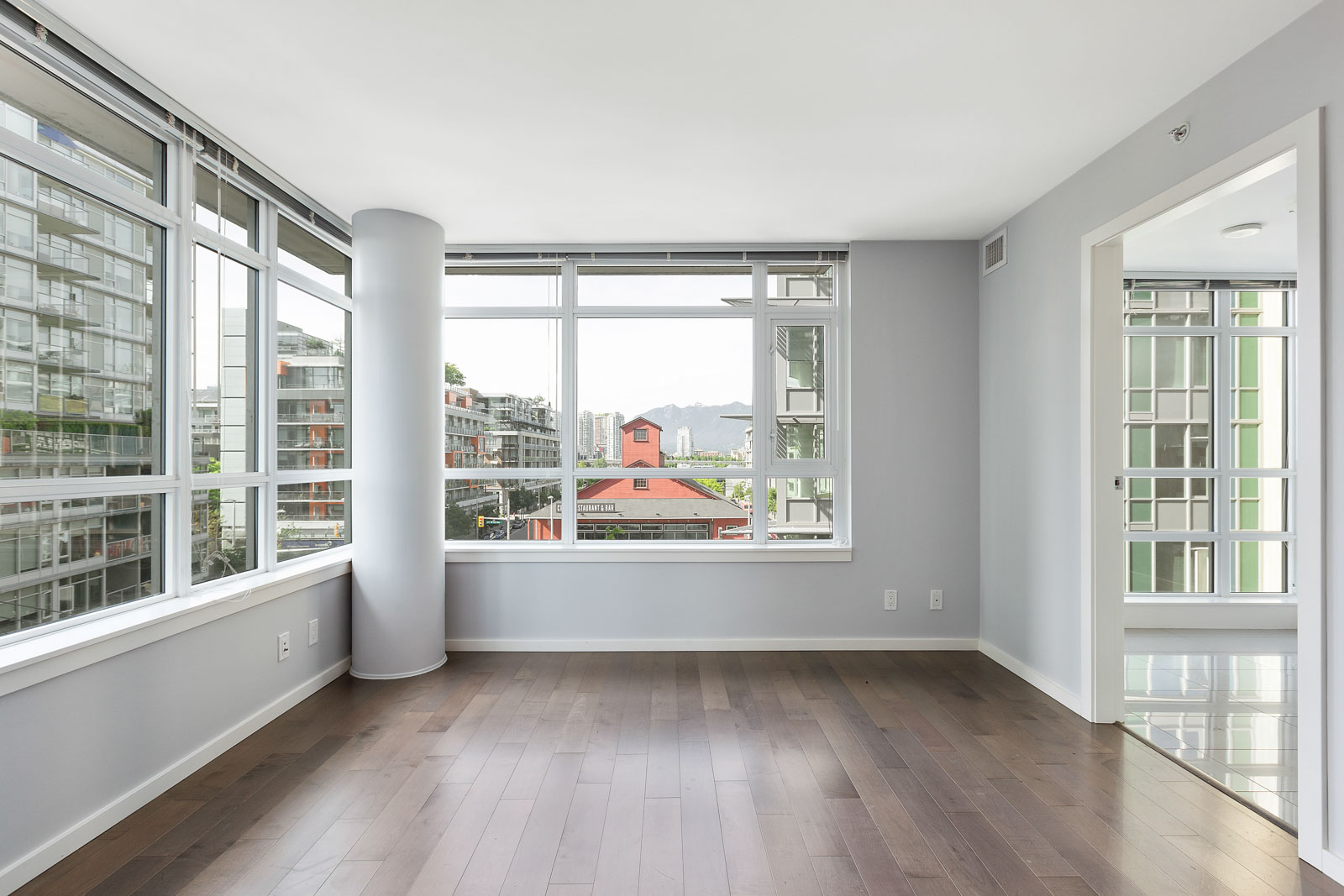Empty modern room with large windows, light gray walls, and dark hardwood floors, offering a city and mountain view.