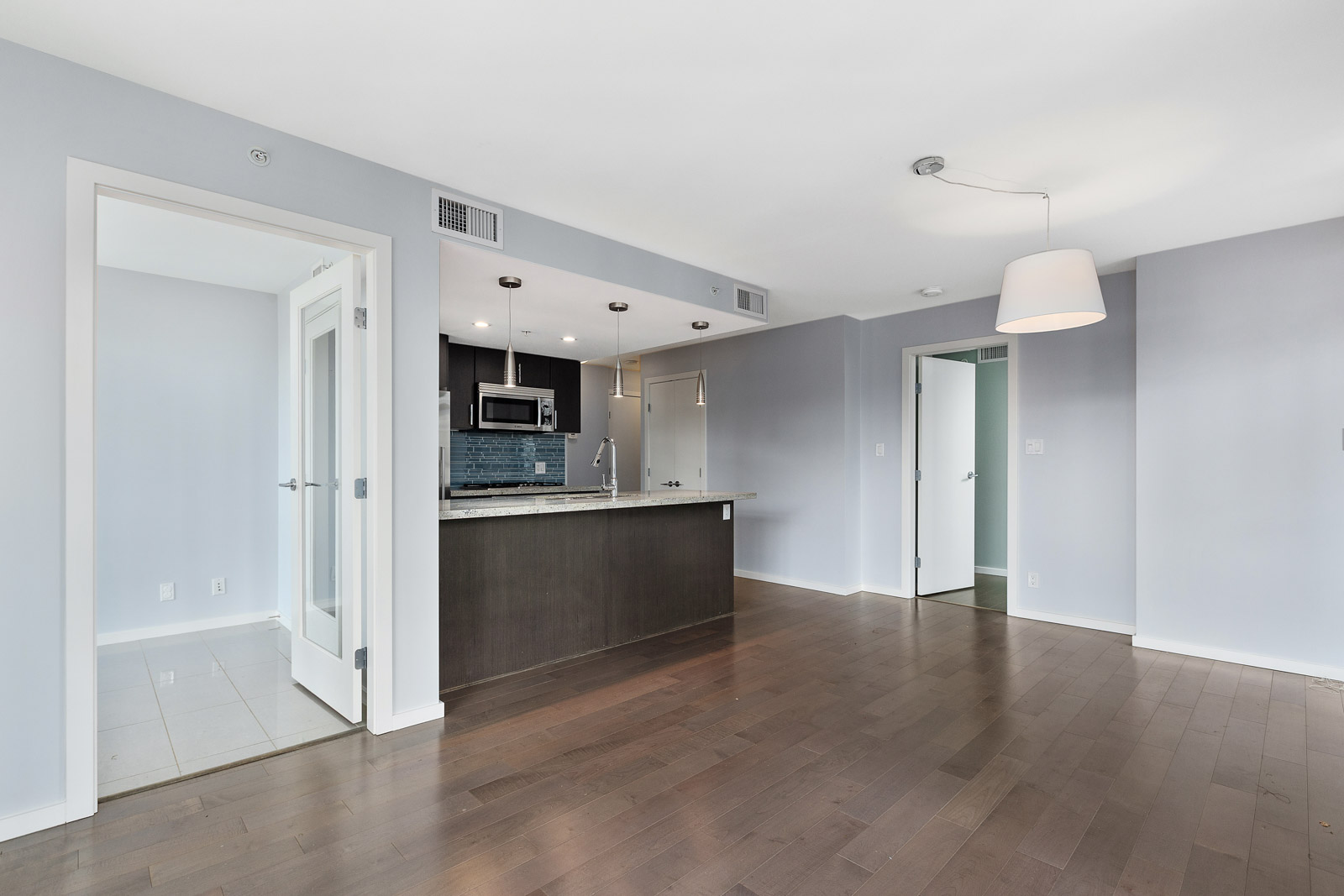Modern apartment interior with hardwood floors, open kitchen featuring dark cabinets and stainless steel appliances, pendant lighting, and adjacent rooms with white doors.