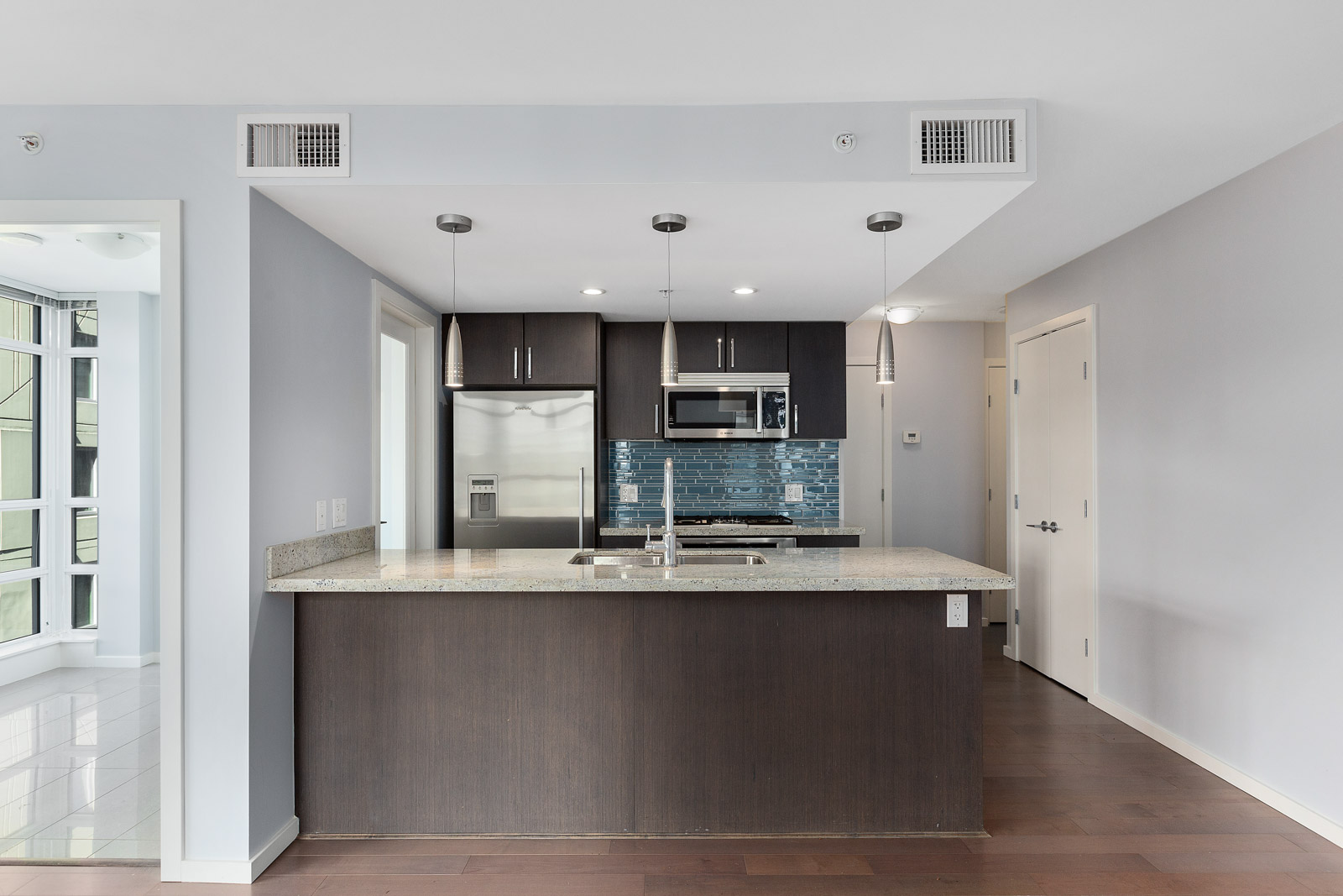 Modern kitchen with dark cabinets, stainless steel appliances, blue subway tile backsplash, island with sink, pendant lights, and hardwood floors.