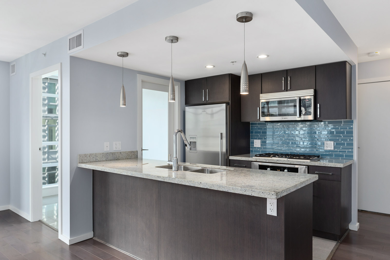 Modern kitchen with dark cabinets, stainless steel appliances, granite countertops, a central island with sink, and blue subway tile backsplash. Three pendant lights hang above the island.