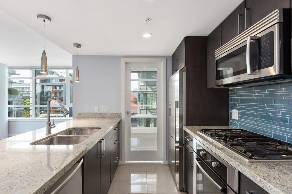Modern kitchen with dark cabinets, granite countertops, stainless steel appliances, gas stove, blue tile backsplash, and large windows providing natural light.