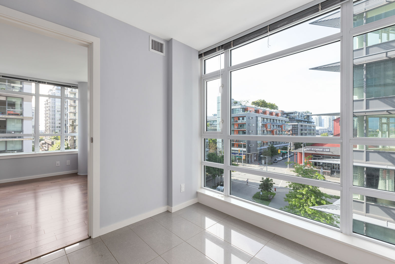 Bright, unfurnished apartment with large floor-to-ceiling windows, tiled floors, and a view of city buildings; adjacent room has wooden flooring.