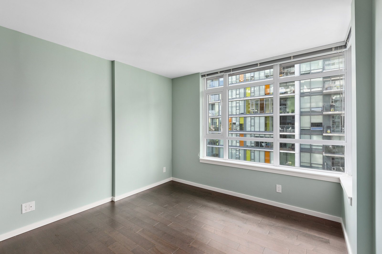 Empty room with light green walls, dark hardwood floor, large window, and view of a modern apartment building exterior.