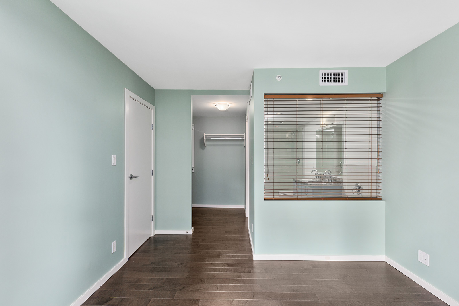 Room with light green walls, dark wood floor, a white closet door, an open closet, and an interior window with blinds facing a bathroom sink and mirror.