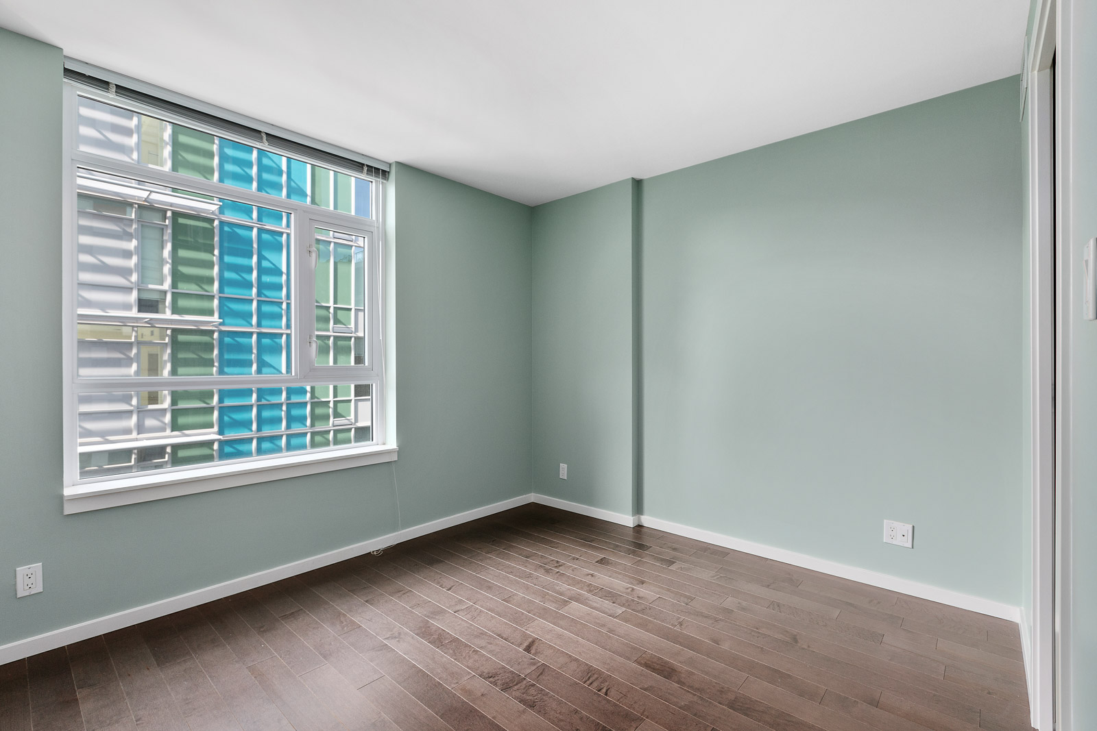 Empty room with light green walls, a large window with a view of a colorful exterior, and dark wood flooring.