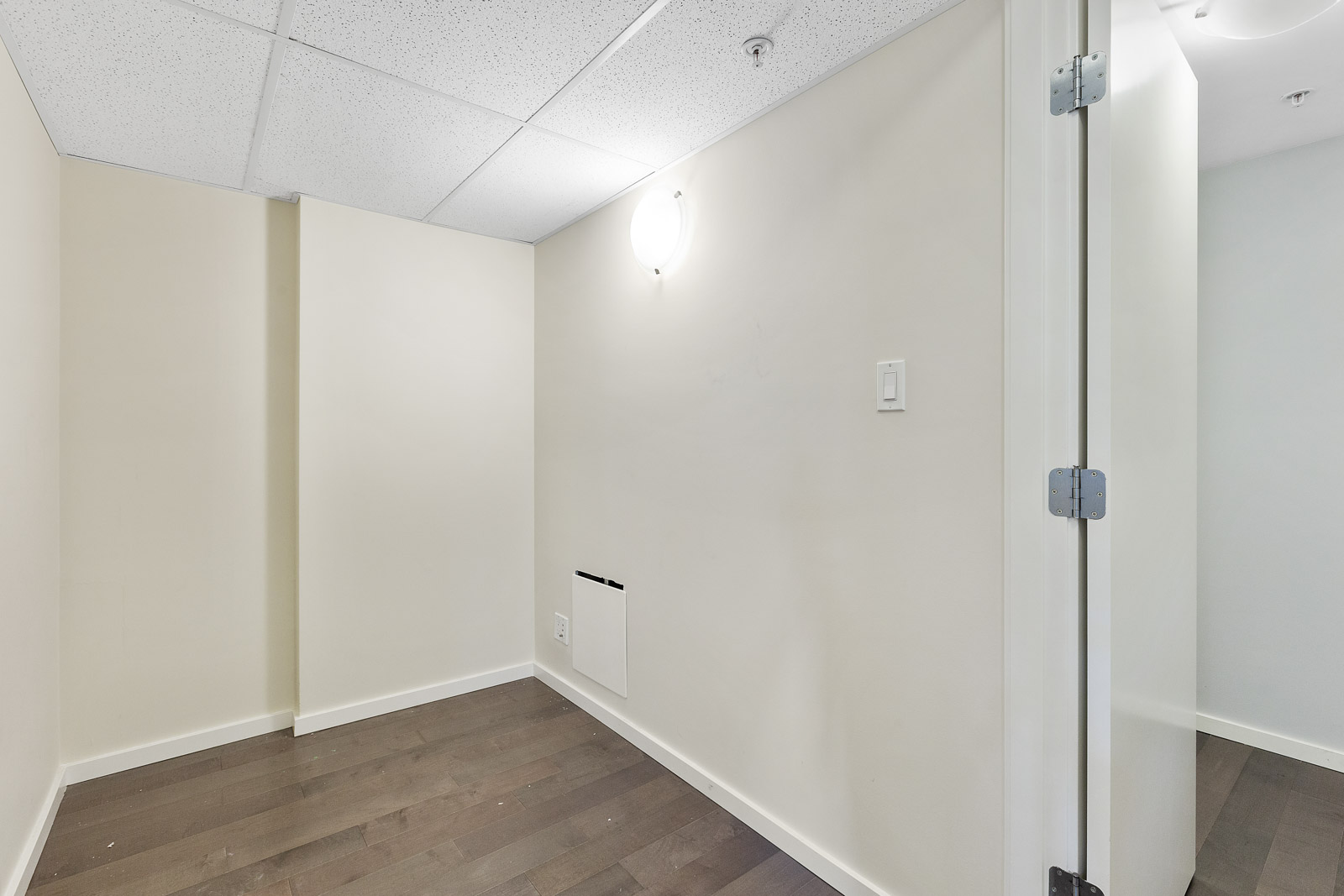 Small, empty room with beige walls, dark hardwood floor, white ceiling tiles, a wall-mounted light fixture, and an open door on the right.