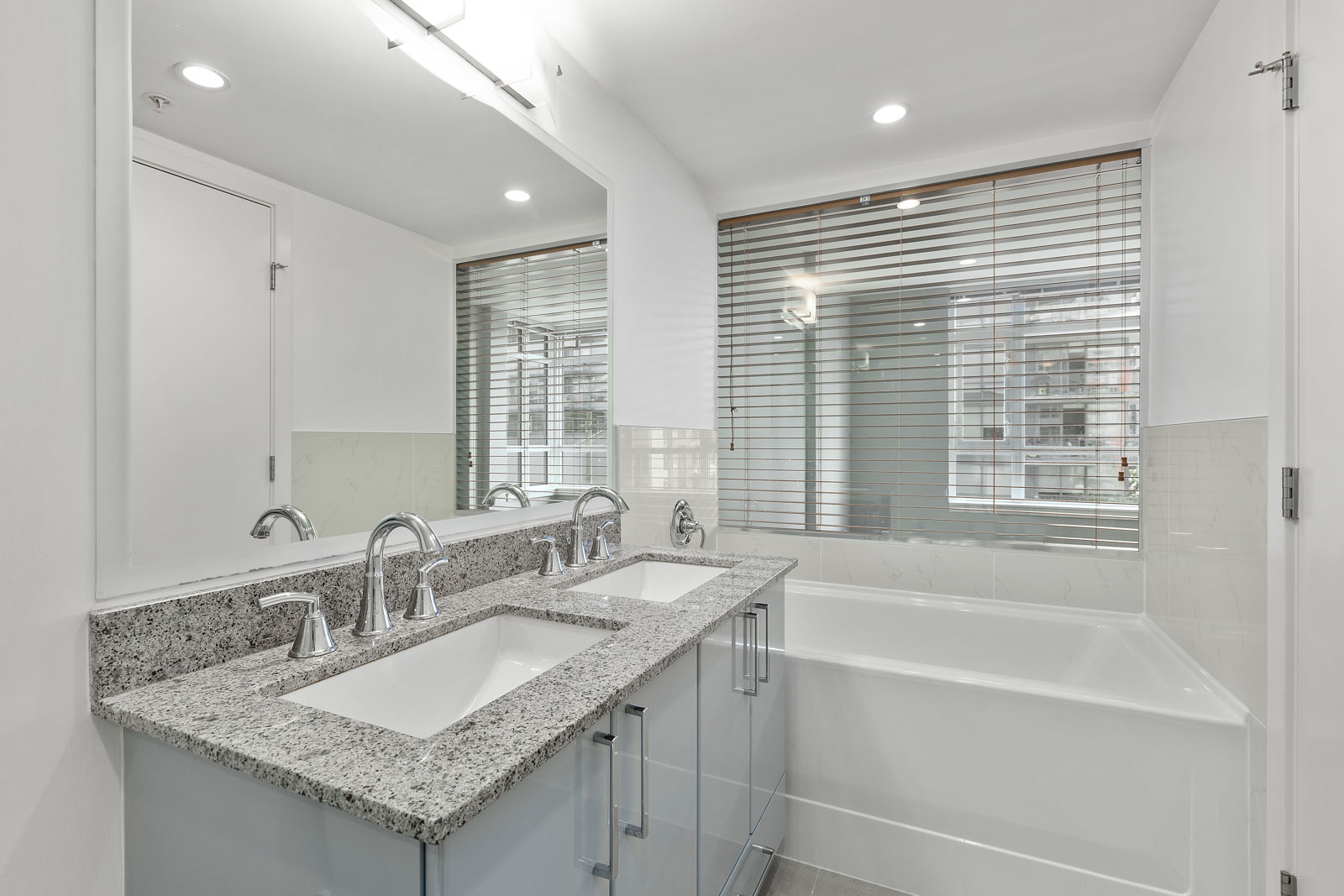 Modern bathroom with double sink vanity, granite countertop, large mirror, built-in bathtub, and window with horizontal blinds.