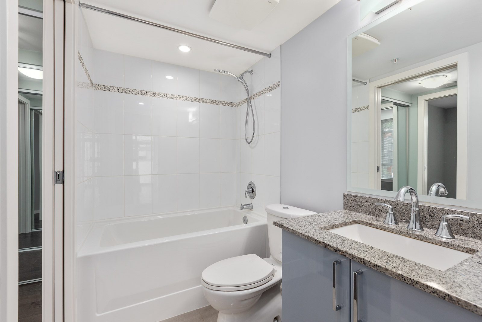 Modern bathroom with white tub-shower combo, wall-mounted showerhead, toilet, and granite countertop vanity with double sinks and mirrors.