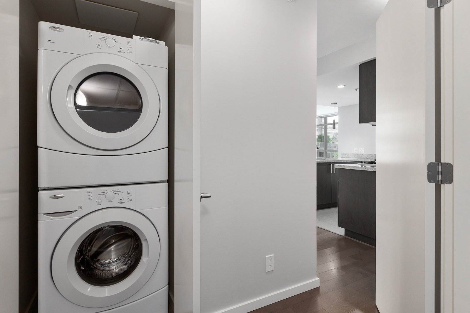 A stacked washer and dryer unit is installed in a small laundry closet adjacent to a hallway leading to a modern kitchen.