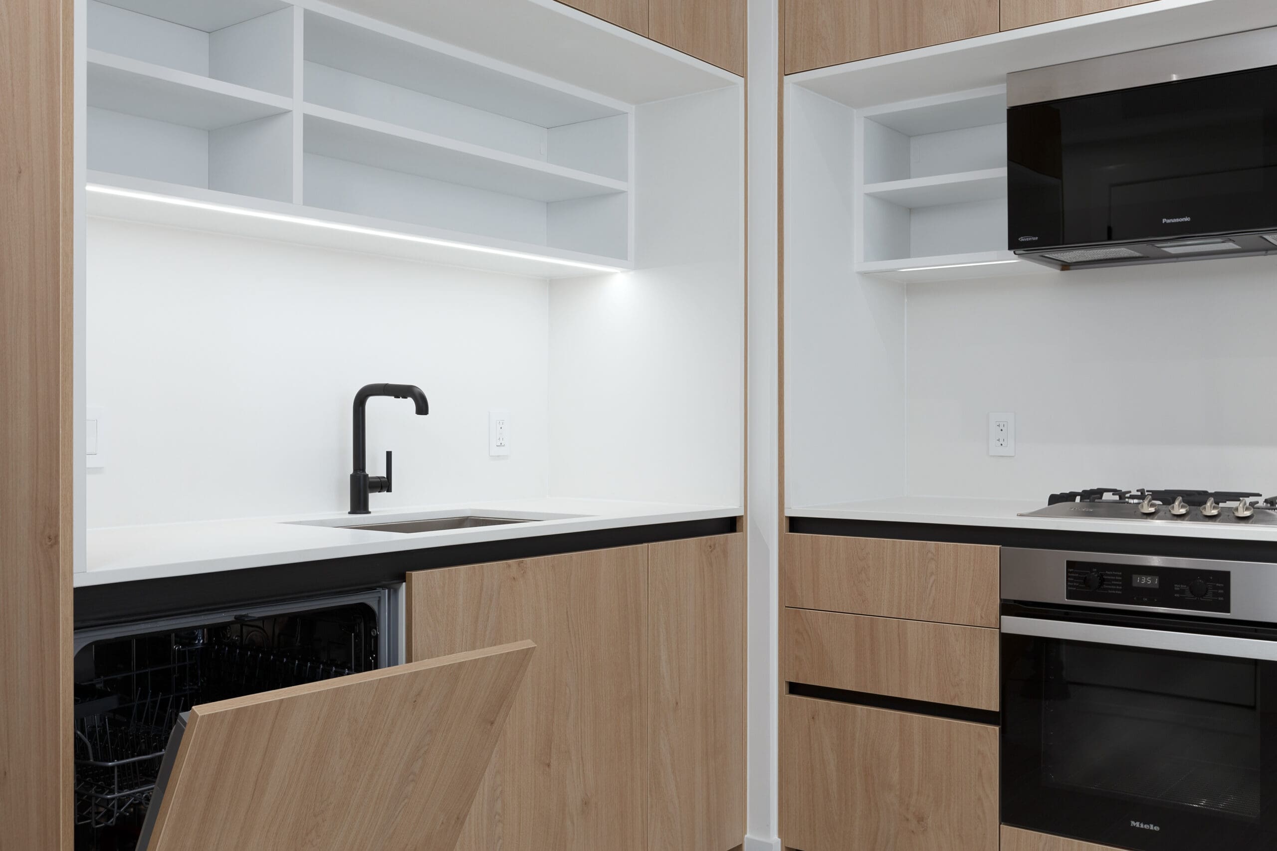 Modern kitchen with light wood cabinets, a built-in oven, gas cooktop, black faucet, open dishwasher, open shelving, and white countertops.