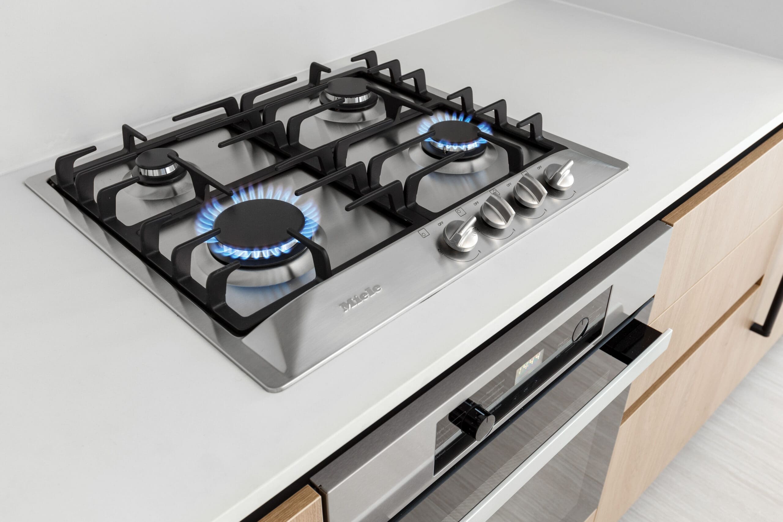 A stainless steel gas cooktop with four burners is turned on, with blue flames visible, set into a modern kitchen countertop above an oven.
