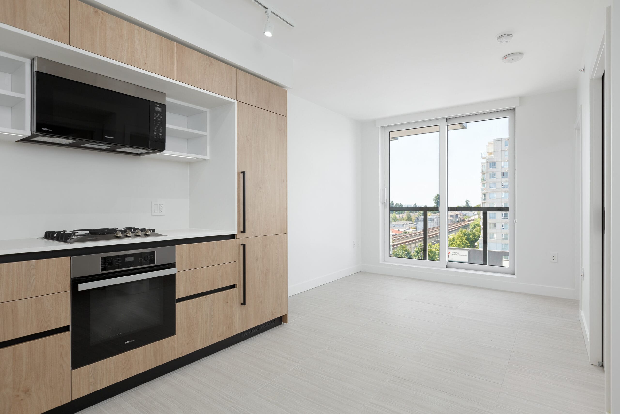 Modern, unfurnished kitchen and dining area with light wood cabinets, built-in oven, gas stove, black microwave, and large window overlooking a cityscape.
