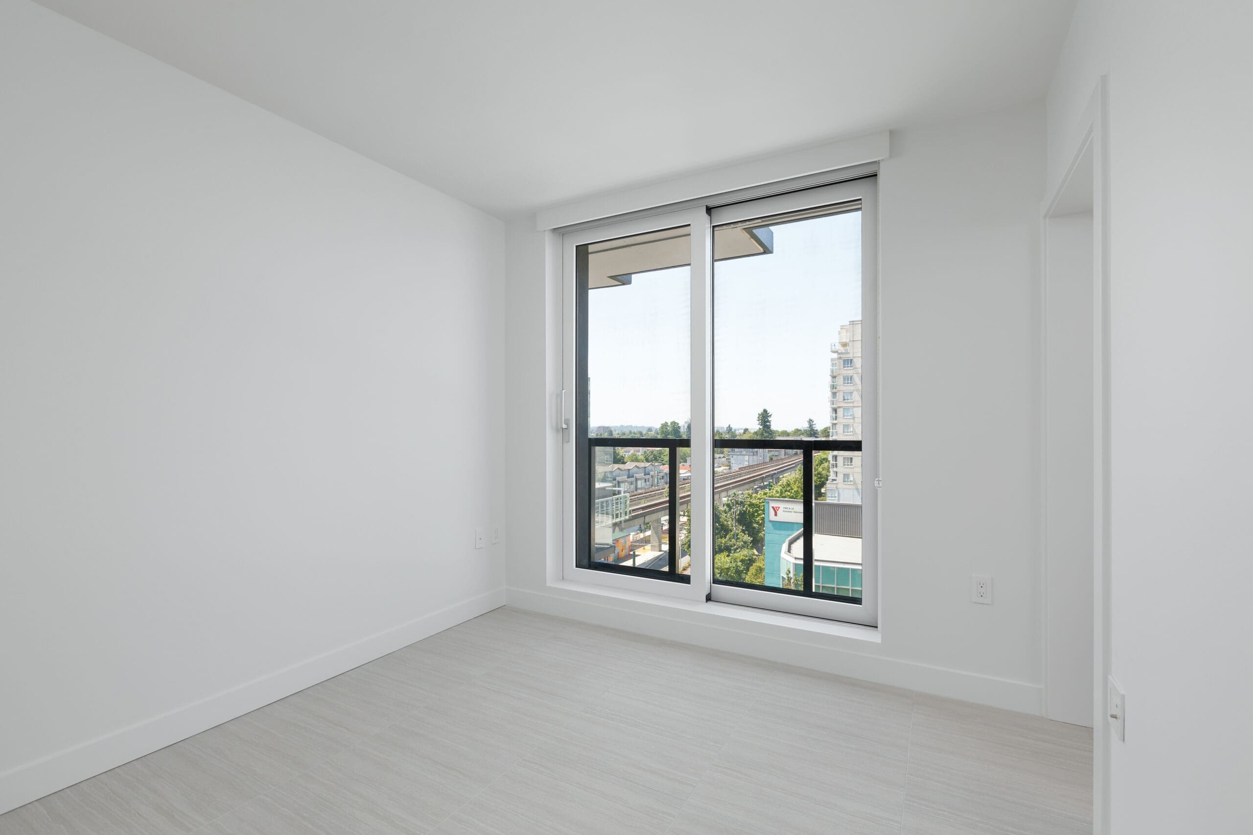 Unfurnished, white-walled room with light wood flooring and large floor-to-ceiling windows overlooking a cityscape.