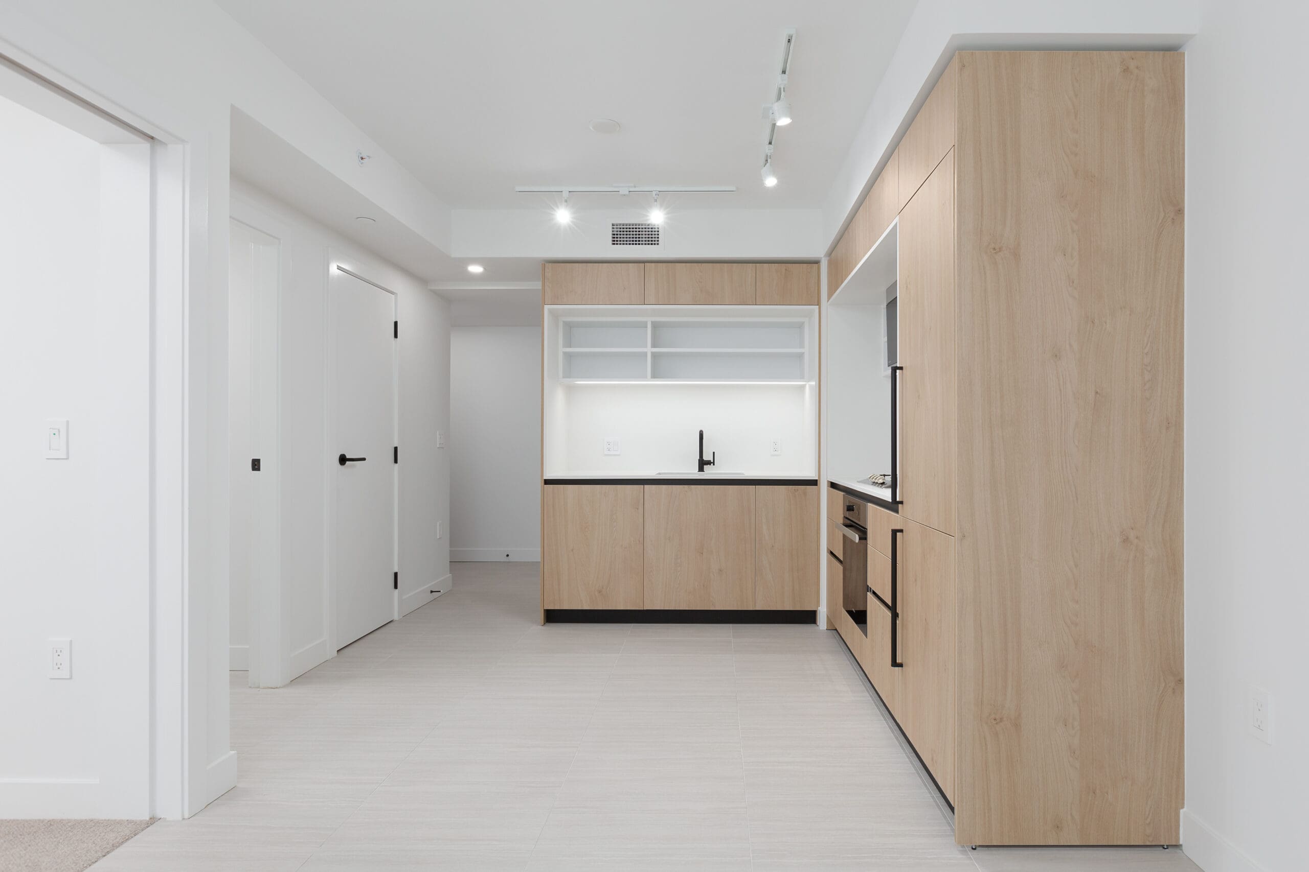 Modern minimalist kitchen with light wood cabinets, white walls, black fixtures, and recessed ceiling lights, adjacent to a hallway with white doors.