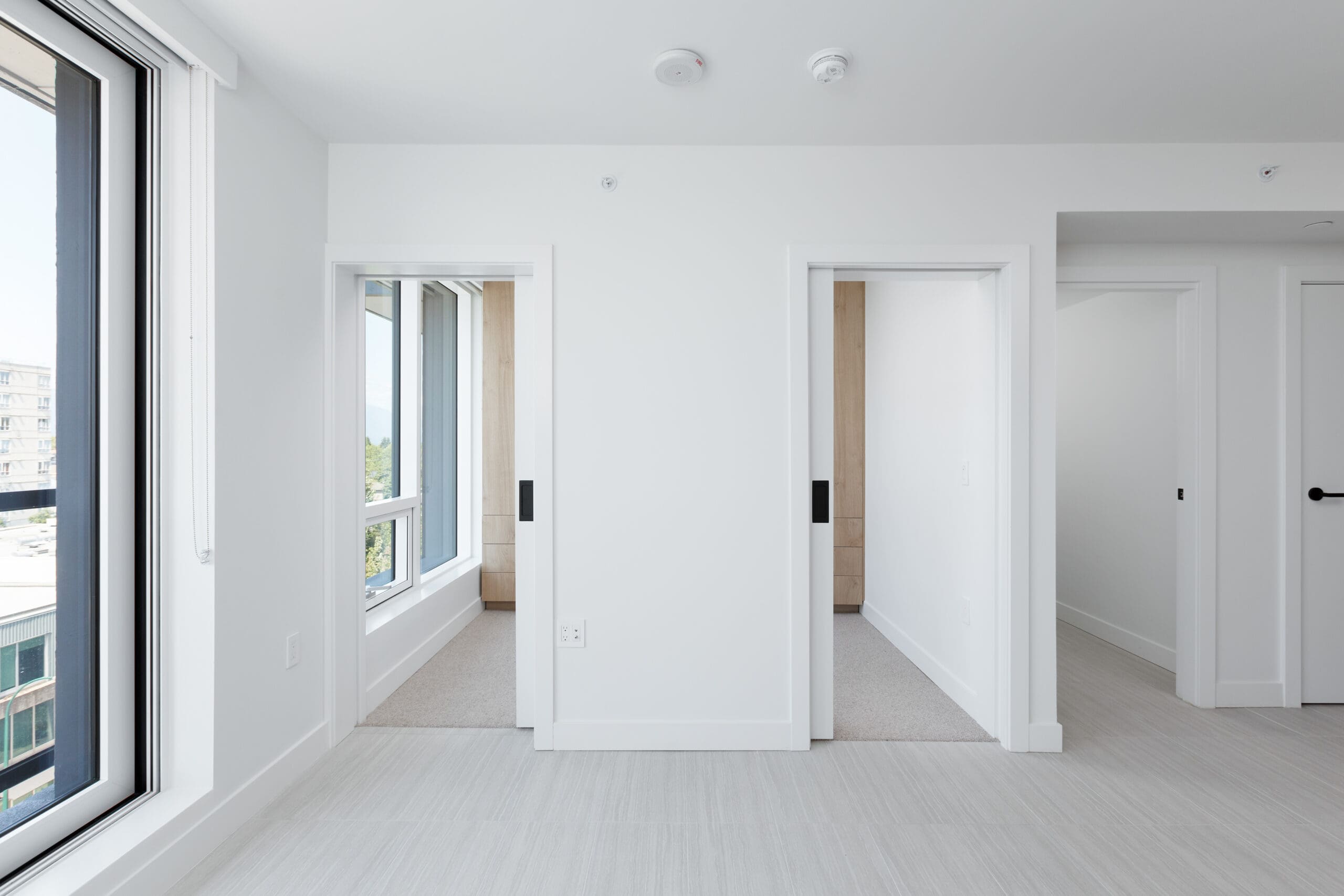 A bright, modern apartment interior with white walls, sliding doors, large windows, and light-colored flooring.
