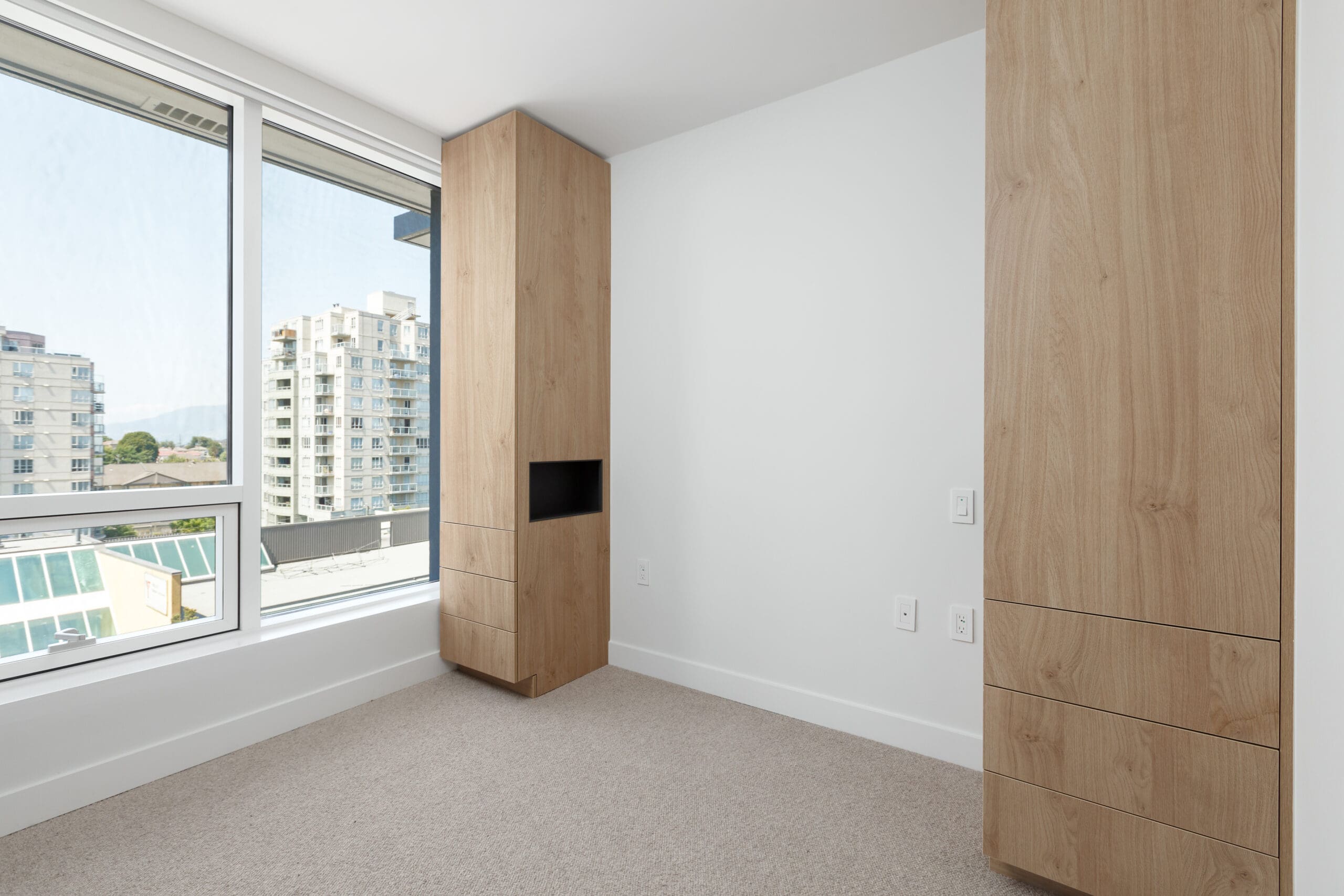 Empty modern room with beige carpet, two tall wooden cabinets, large window with city view, and white walls.
