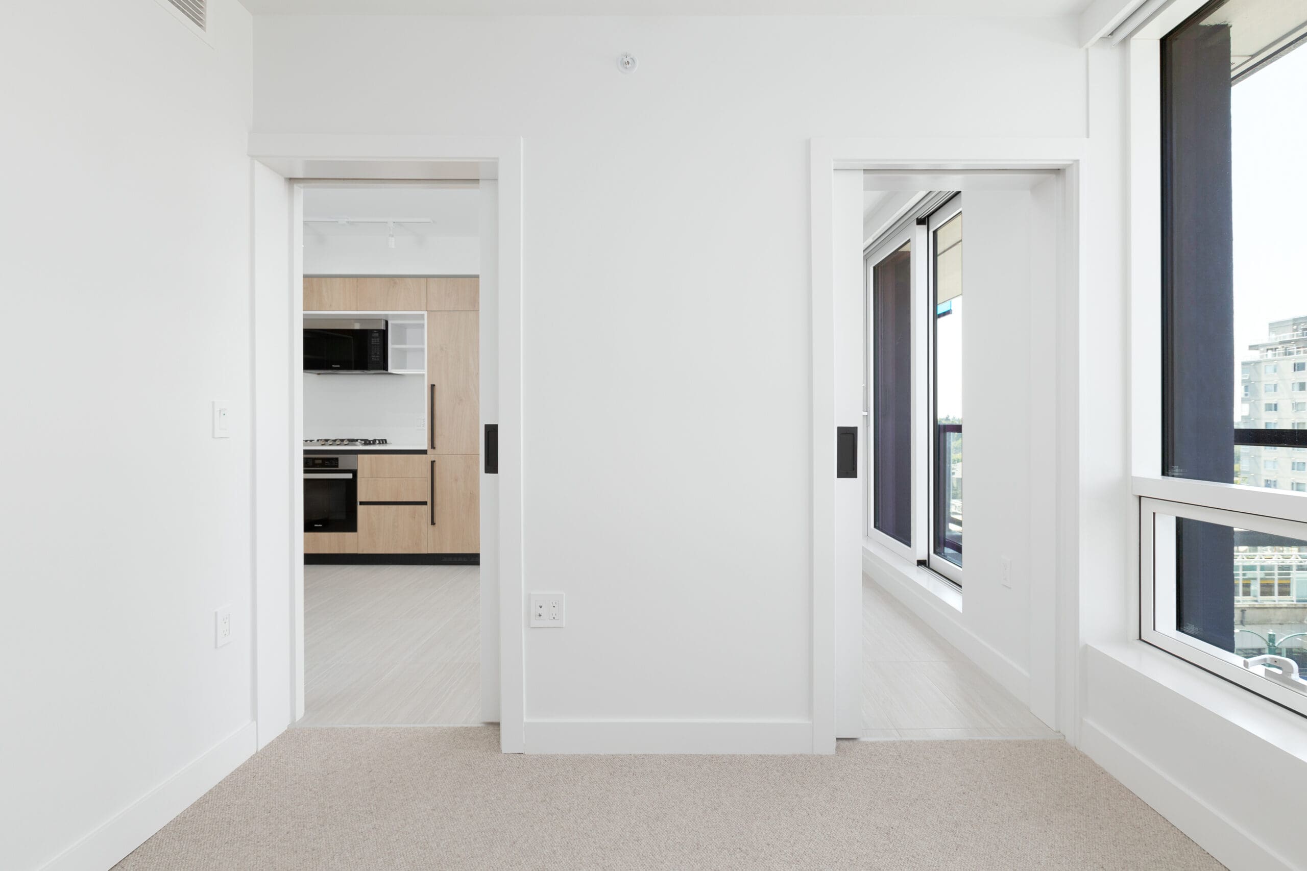 A modern apartment interior with two sliding doors; one leading to a kitchen with light wood cabinets, and the other to a bright room with large windows.