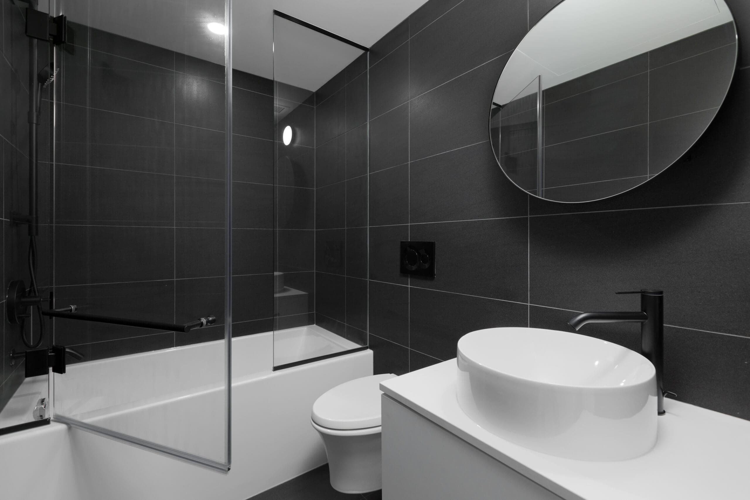 Modern bathroom with dark tiled walls, a glass shower enclosure over a bathtub, a round mirror, and a white vanity with a vessel sink and black fixtures.