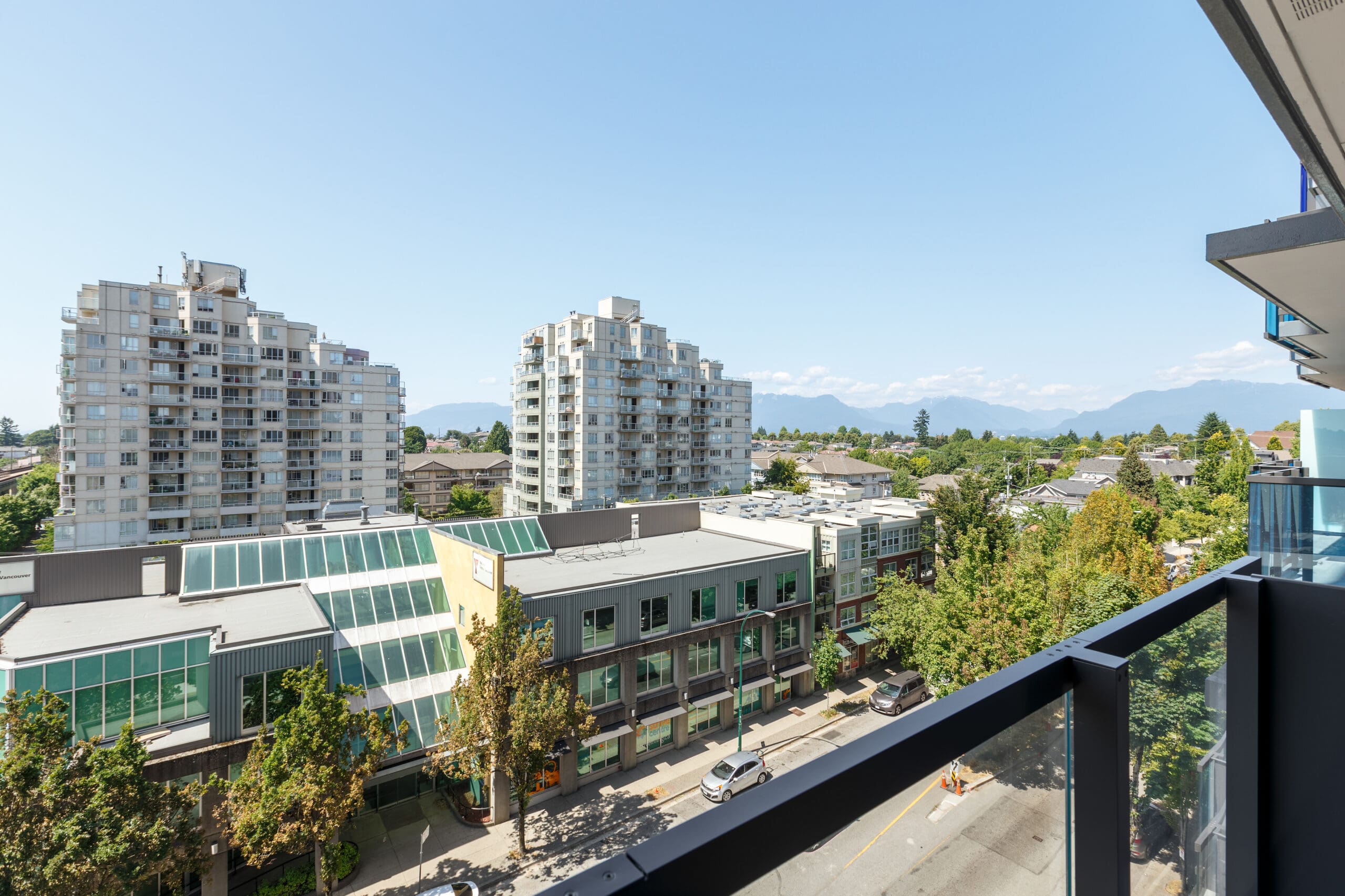 View from a balcony overlooking a city street, with mid-rise apartment buildings, trees, and mountains visible in the background under a clear blue sky.