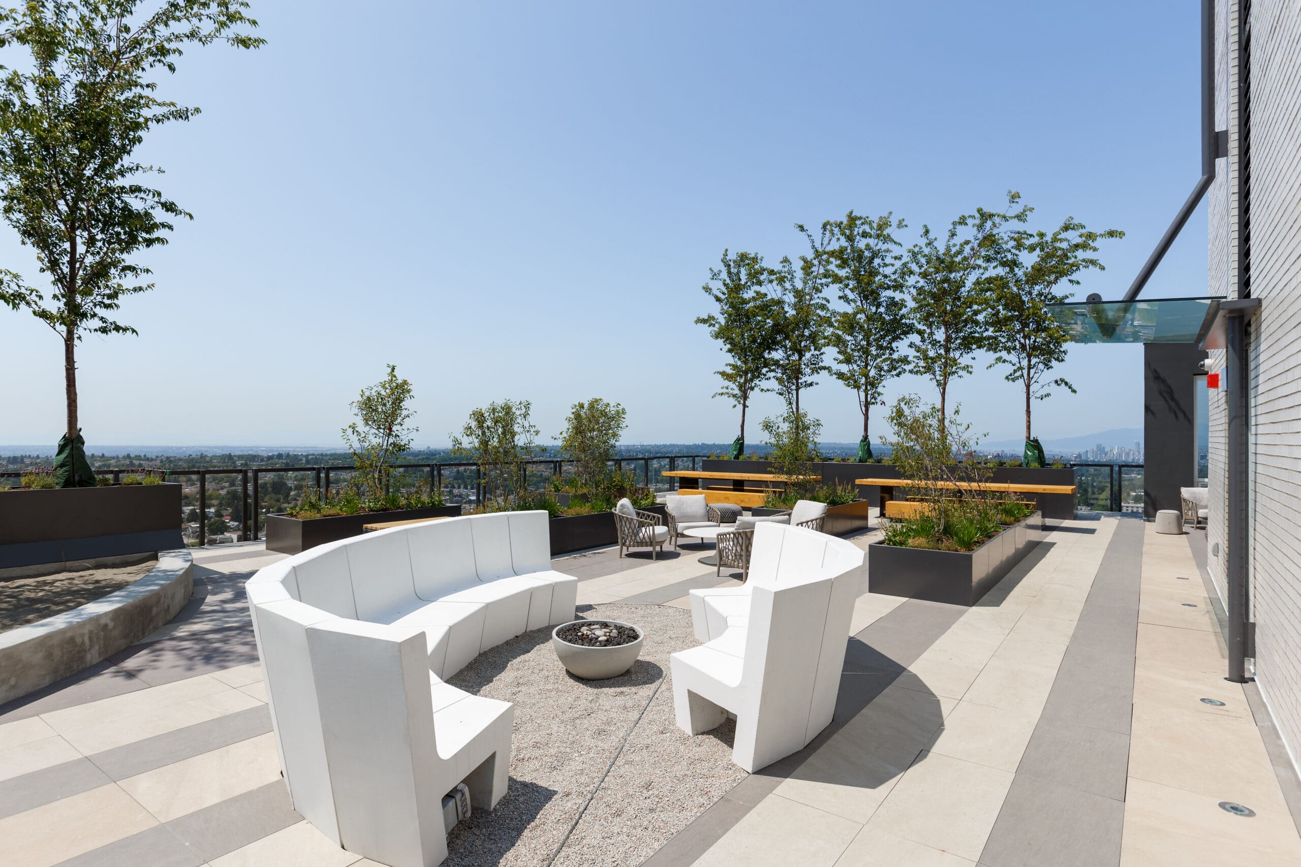 Modern rooftop patio with white curved seating around a fire pit, trees in planters, and a city view in the background on a sunny day.