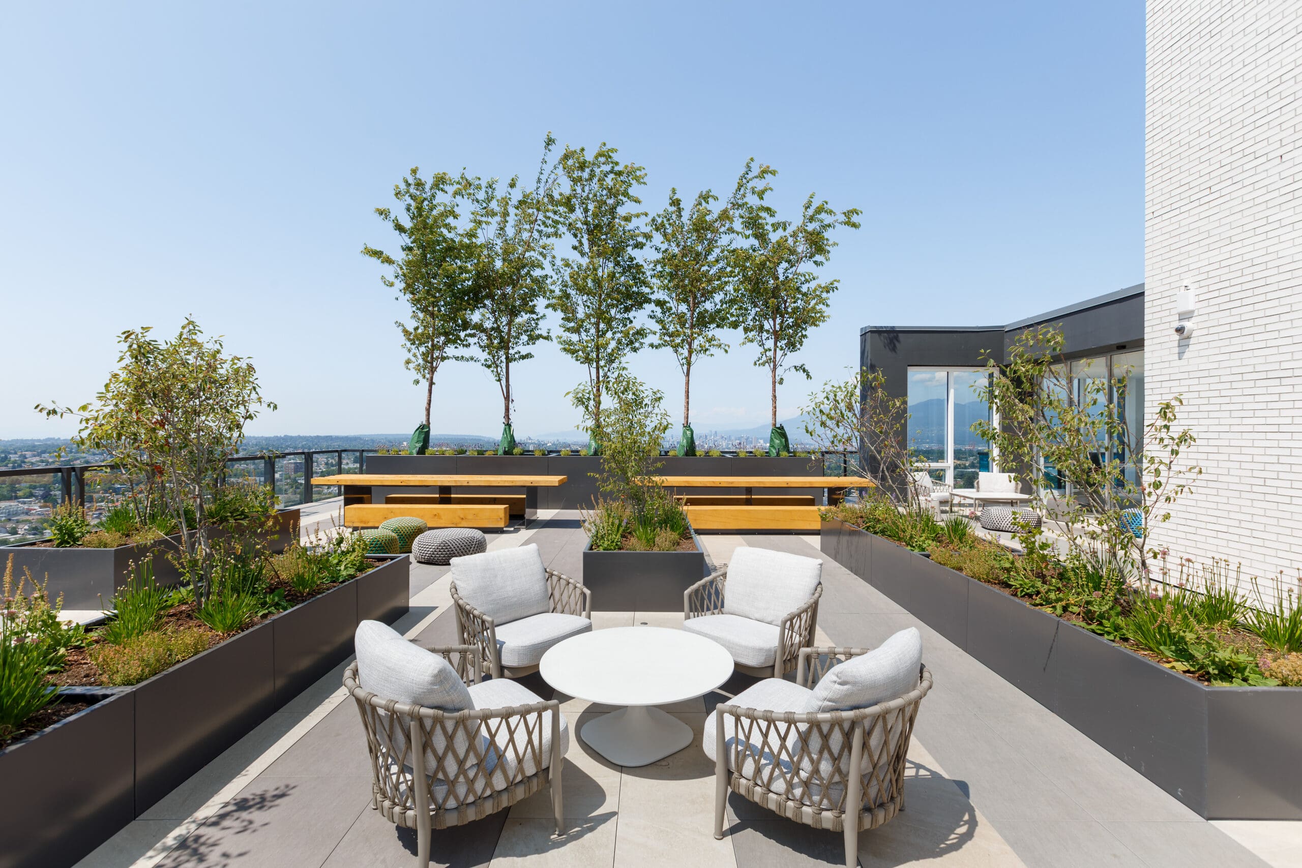 Rooftop patio with four wicker chairs around a white table, planter boxes with greenery, benches, and a city view in the background.