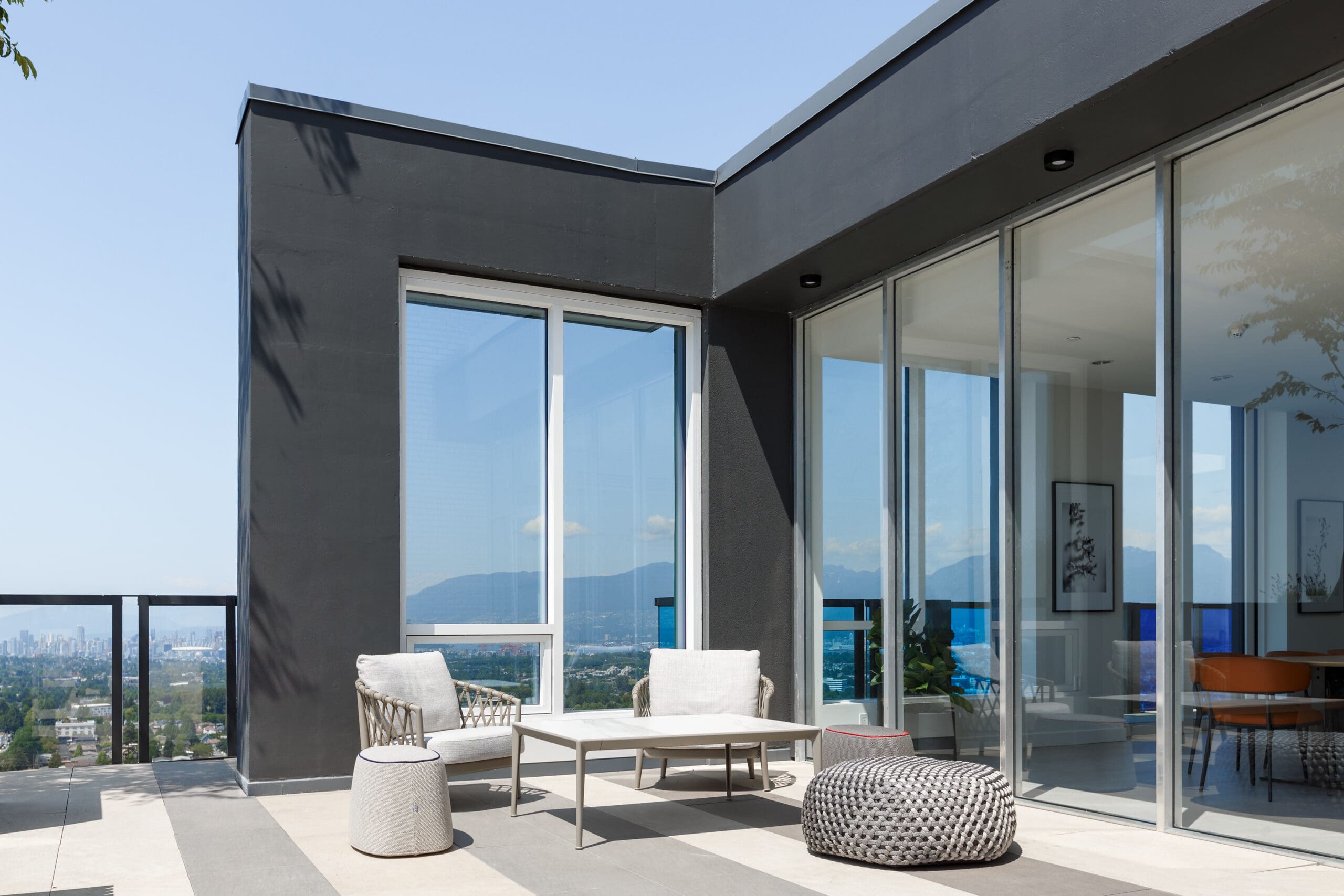 Modern patio with a lounge chair, table, and pouf next to large glass windows, overlooking a cityscape and mountains in the distance.