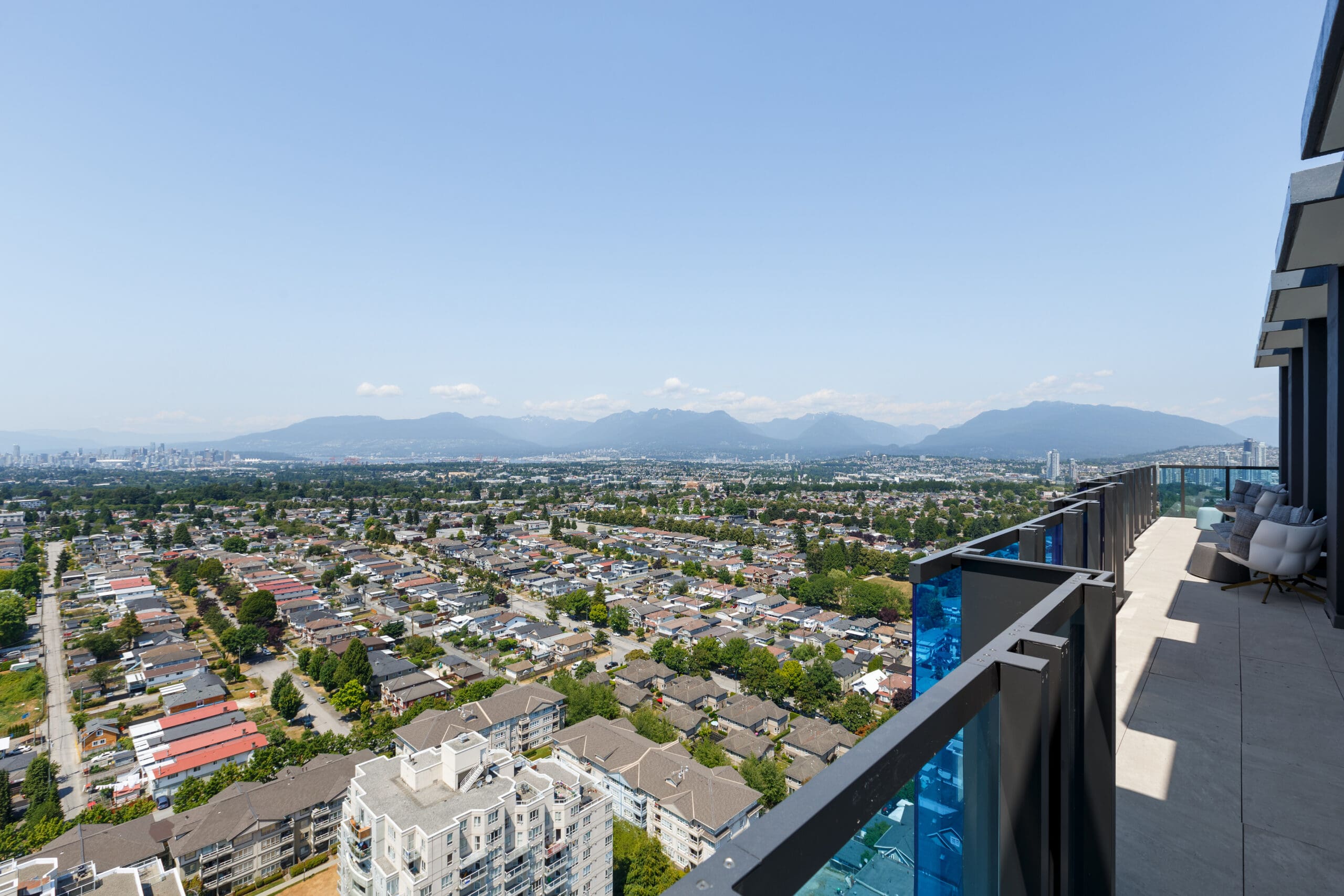 High-rise balcony view overlooking a cityscape with residential buildings, greenery, and distant mountains under a clear blue sky.