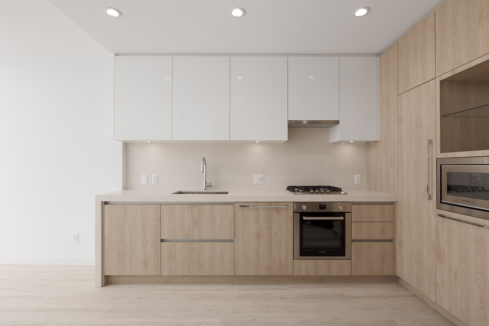 Modern kitchen with light wood lower cabinets, white upper cabinets, built-in oven, cooktop, sink, and integrated microwave set against a minimalist white wall.