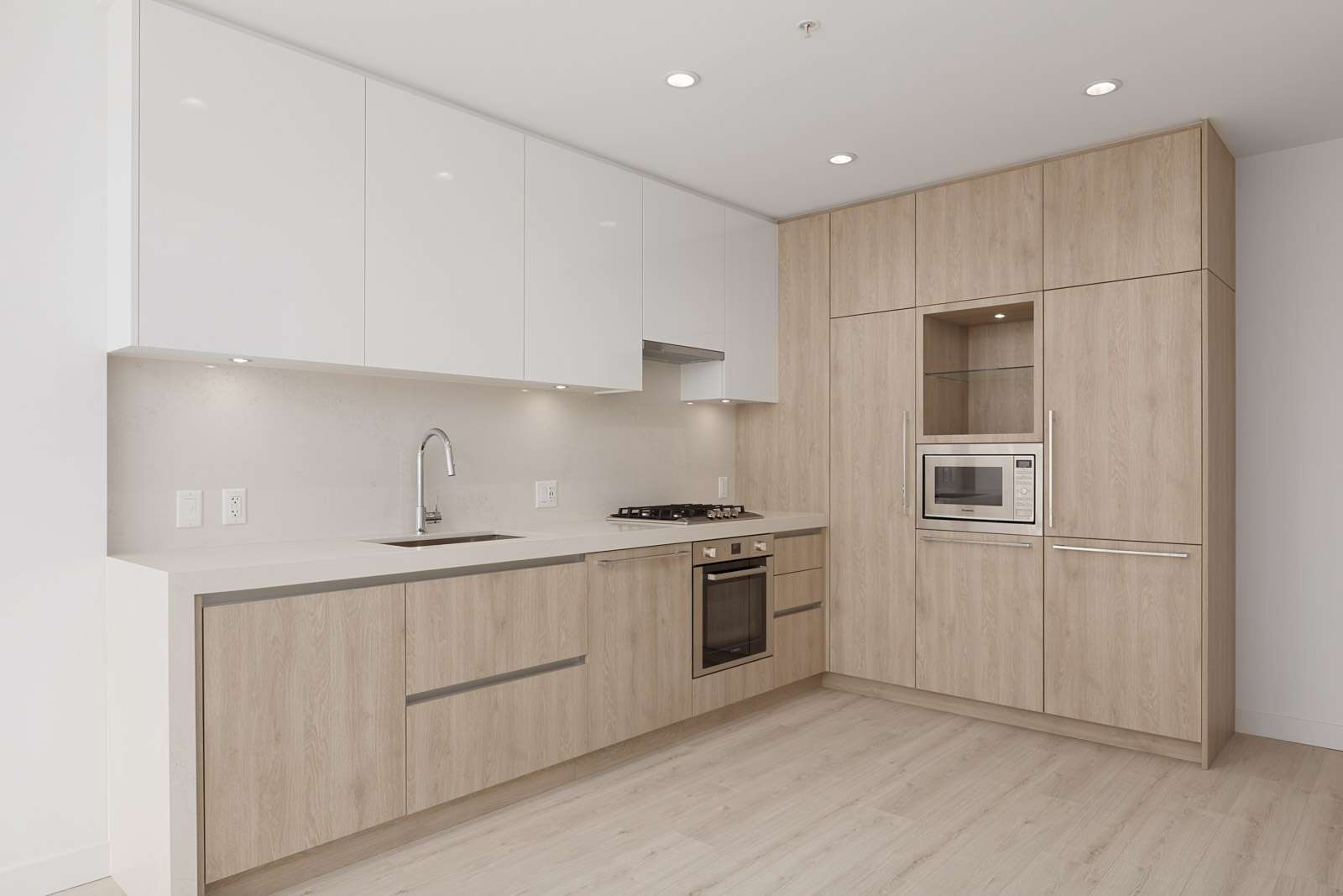Modern kitchen with light wood cabinets, white upper cabinets, built-in oven, stovetop, microwave, and under-cabinet lighting on a light wood floor.