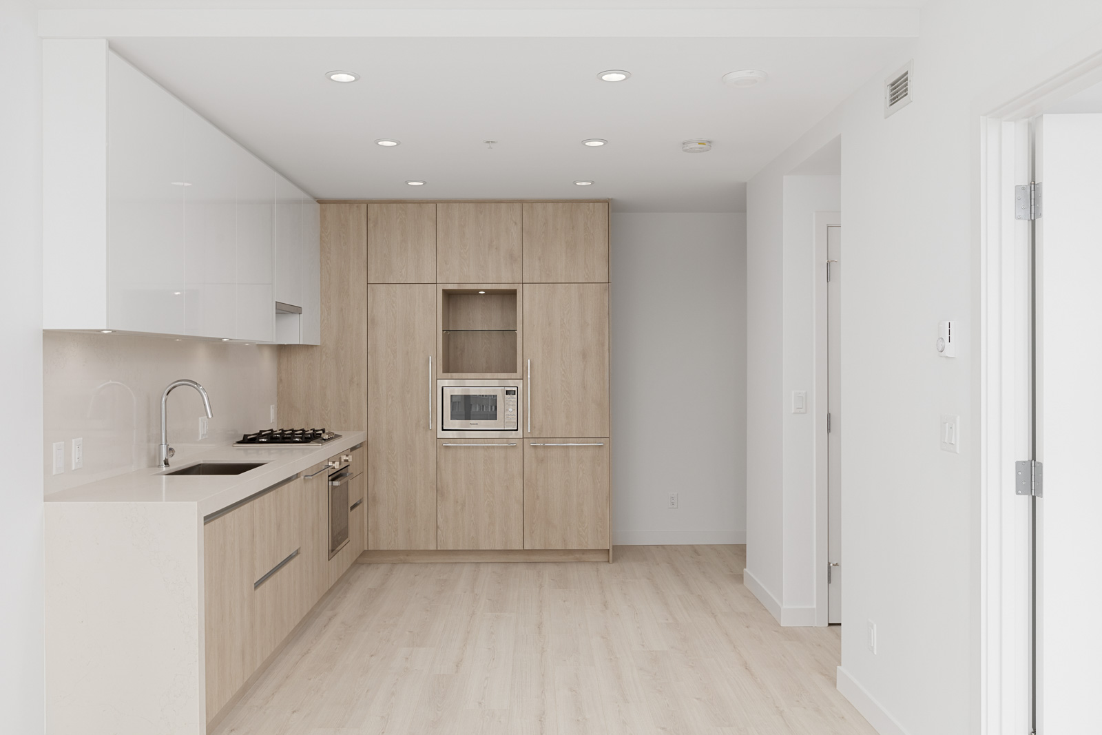 Modern kitchen with light wood cabinets, built-in oven and microwave, white countertops, and integrated sink under recessed lighting in a minimal apartment.