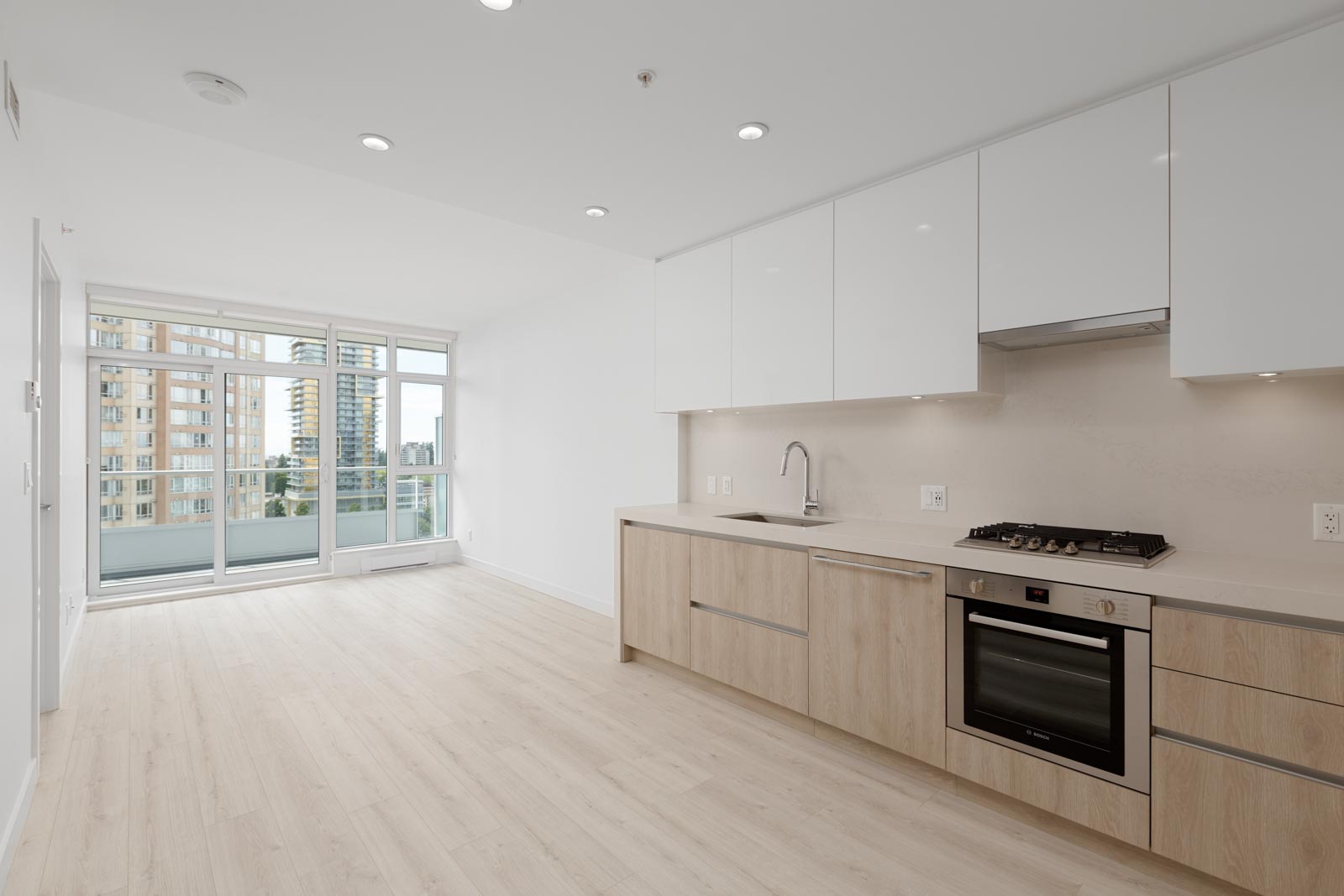 Modern, unfurnished apartment with light wood flooring, white cabinets, built-in oven, gas cooktop, and large windows overlooking city buildings.