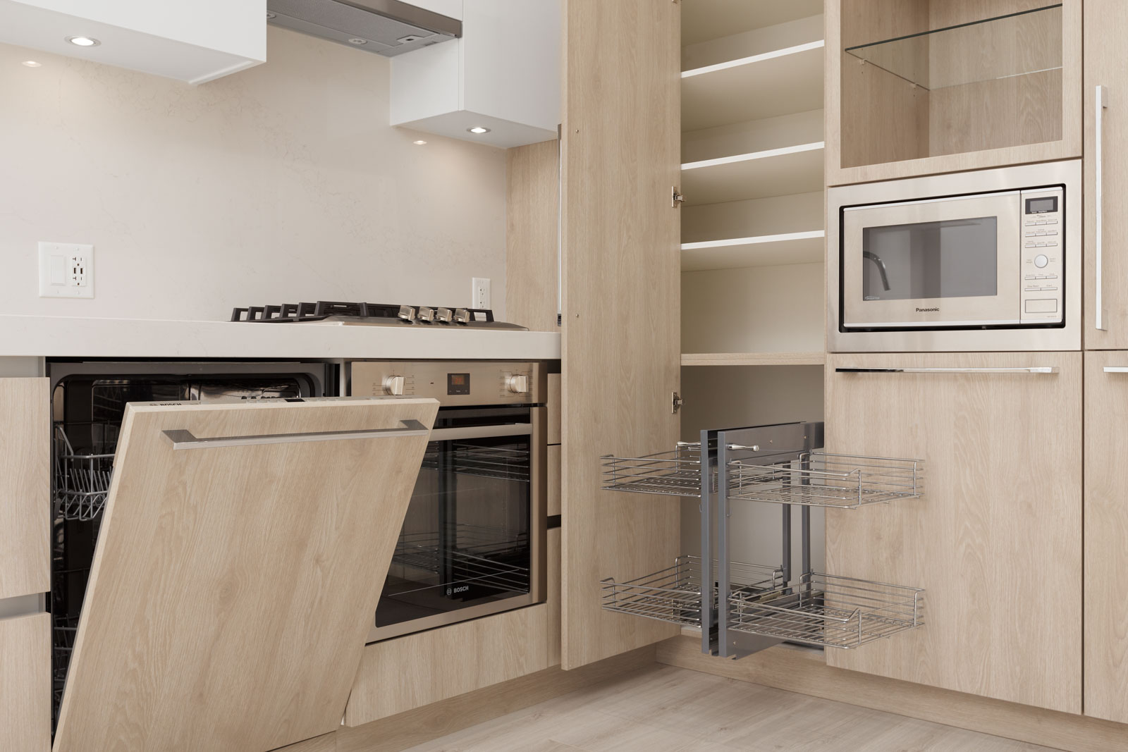 Modern kitchen with light wood cabinetry, open dishwasher, pull-out wire shelving, built-in oven, gas cooktop, and microwave in a cabinet next to shelves.