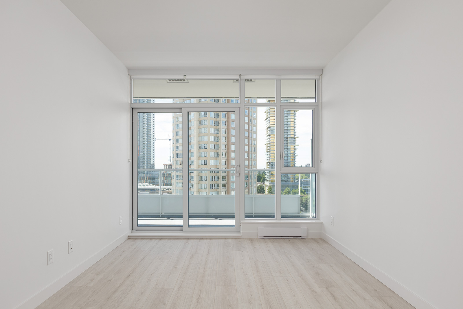 Unfurnished, empty room with light wood flooring, white walls, and large floor-to-ceiling windows opening to a balcony overlooking city buildings.