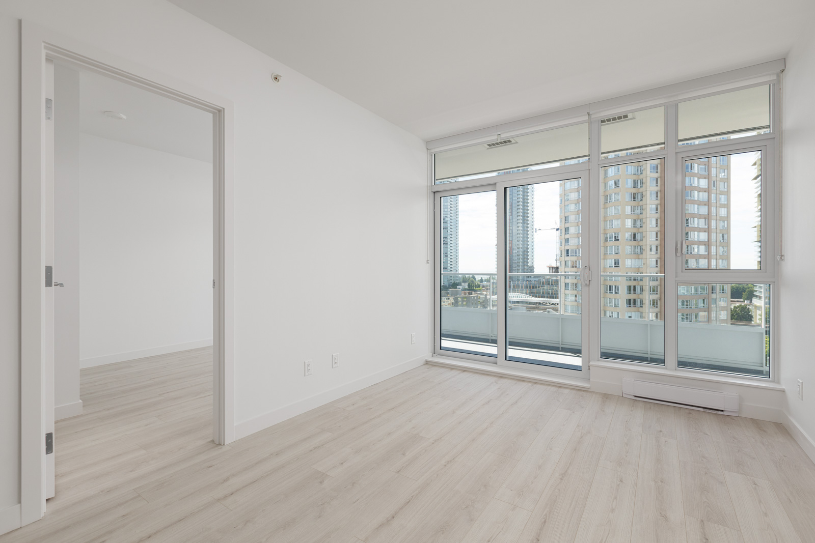 Empty modern apartment room with light wood floors, large windows, glass balcony door, and view of nearby tall buildings. Open doorway leads to another room.