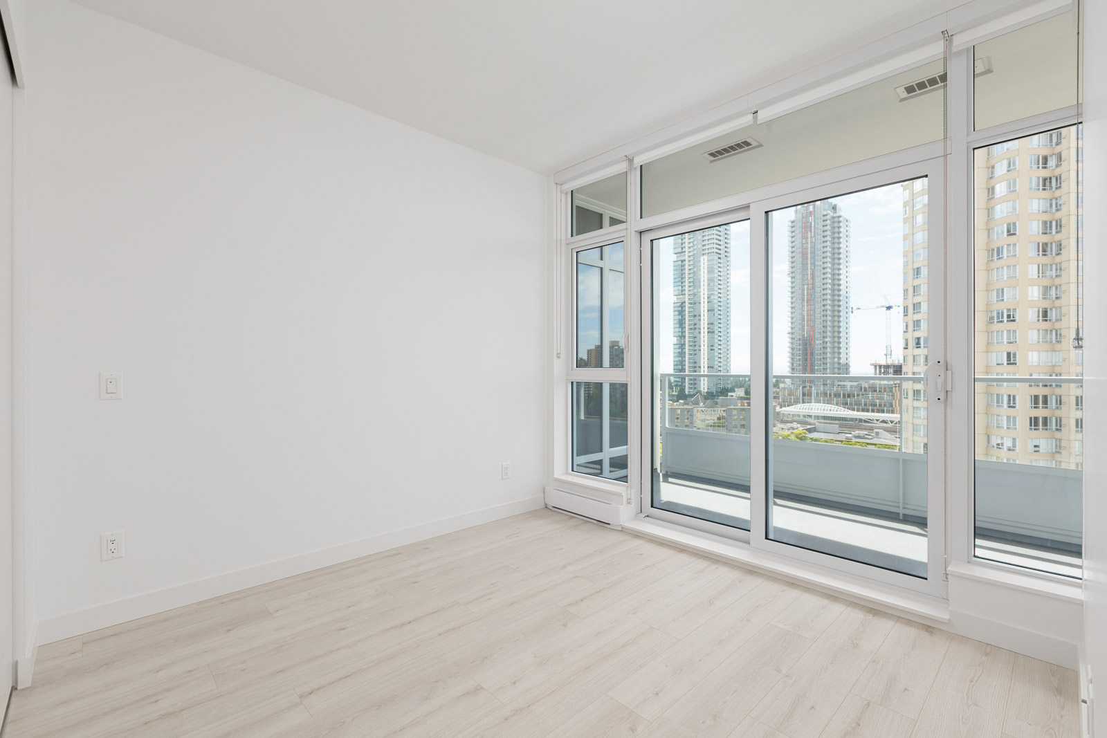 Empty, modern room with light wood flooring, white walls, large windows, and sliding glass doors leading to a balcony overlooking tall city buildings.