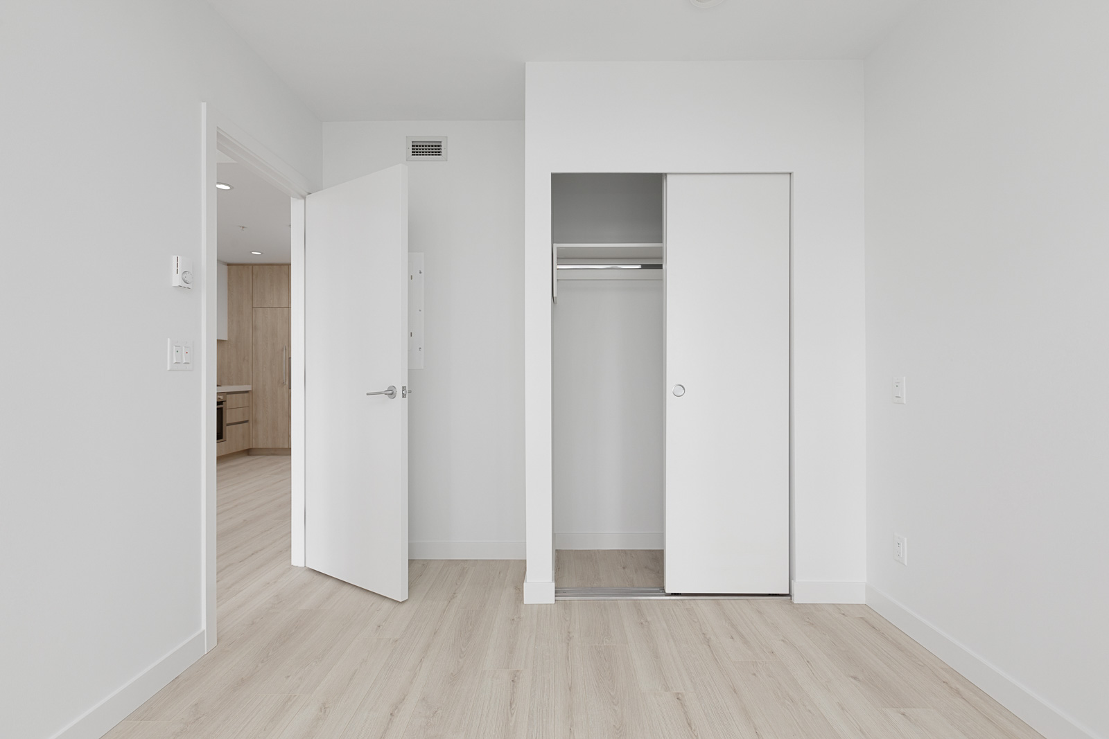 Empty, white-walled room with light wood floor, open door, and closet with sliding doors. Adjoining room with similar decor visible through doorway.
