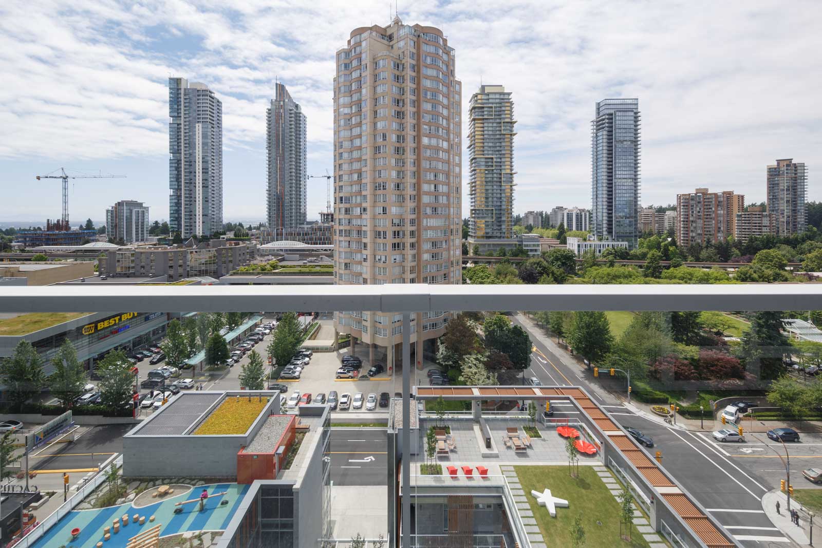 View from a high balcony overlooking a cityscape with tall apartment buildings, green rooftops, a street, parked cars, and a grassy area with benches and a white sculpture.