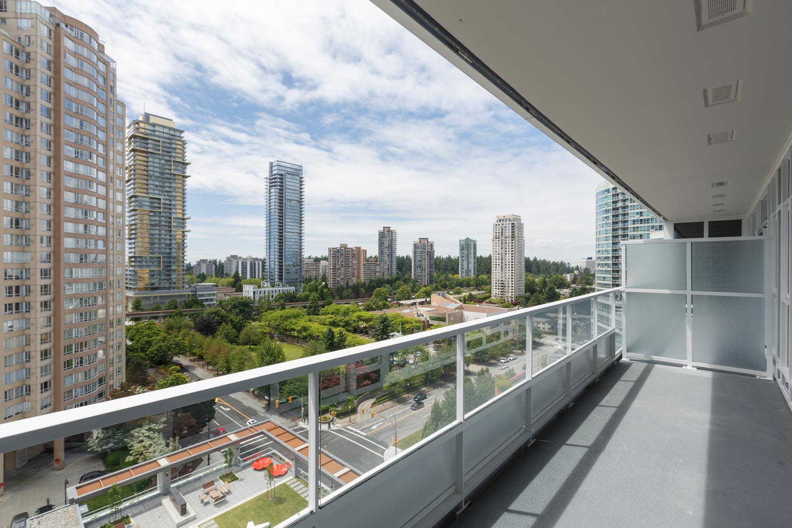 View from a high-rise balcony overlooking a cityscape with tall buildings, green park areas, and a partly cloudy sky.