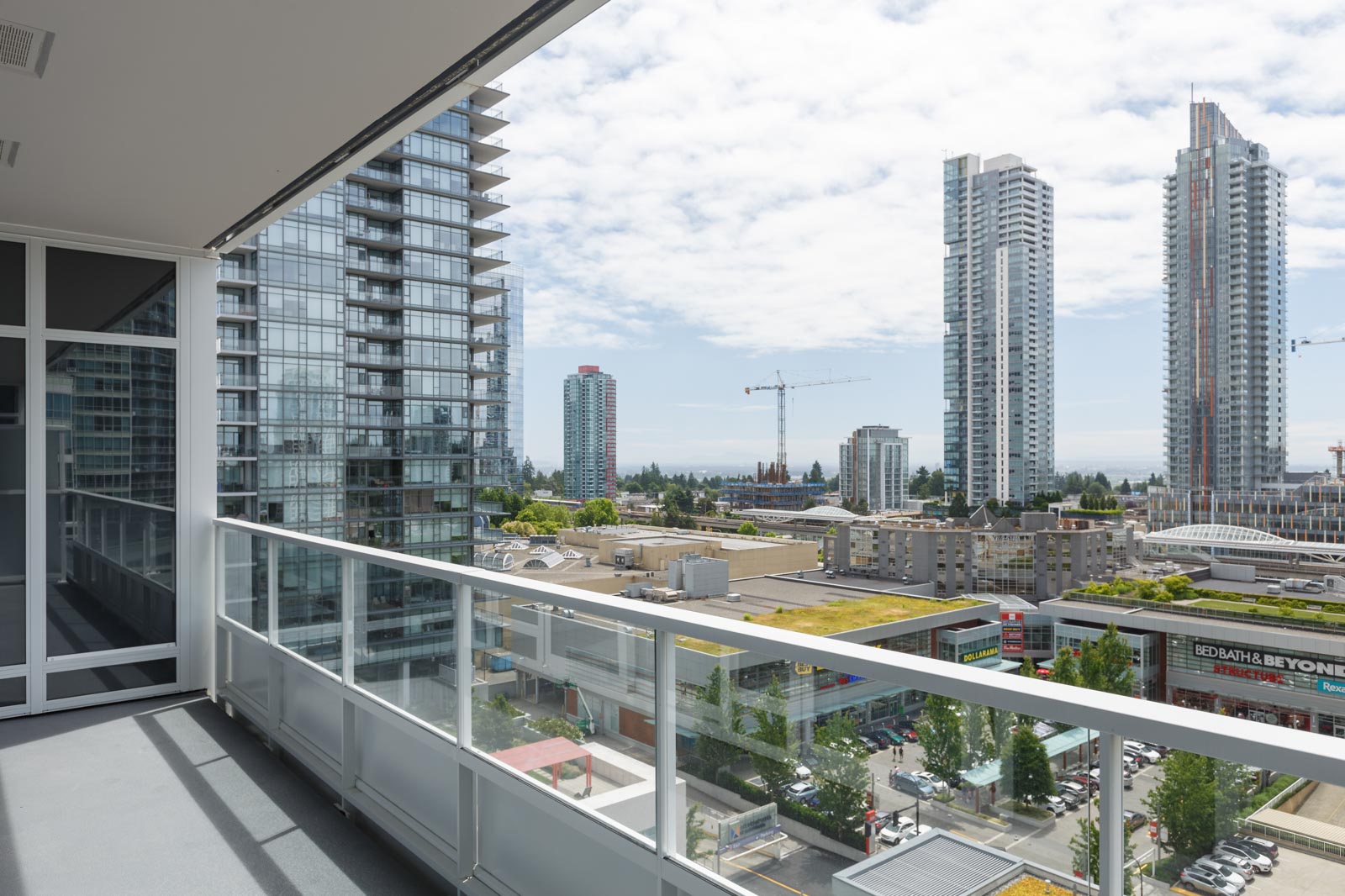 View from a high-rise balcony overlooking city buildings, glass towers, construction cranes, and a partly cloudy sky.