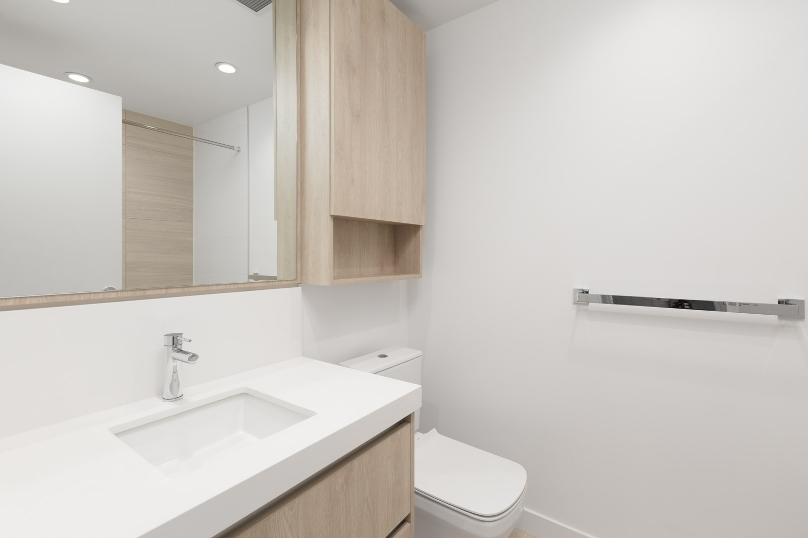 Minimalist bathroom with light wood cabinets, a white countertop and sink, a wall mirror, toilet, towel bar, and plain white walls.