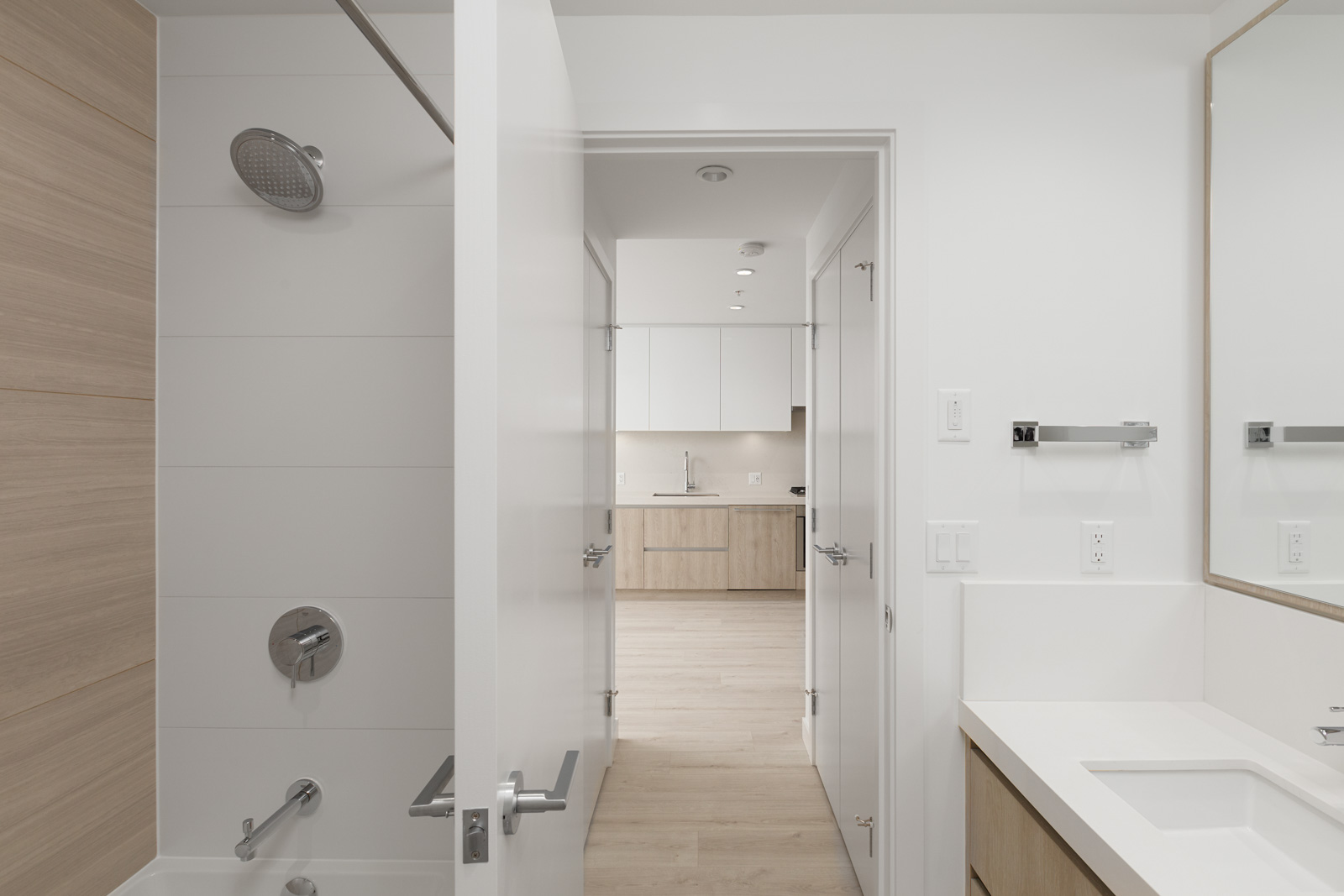 Modern bathroom with a bathtub, shower, and sink, viewed through an open door leading to a bright kitchen area with wood and white cabinetry.